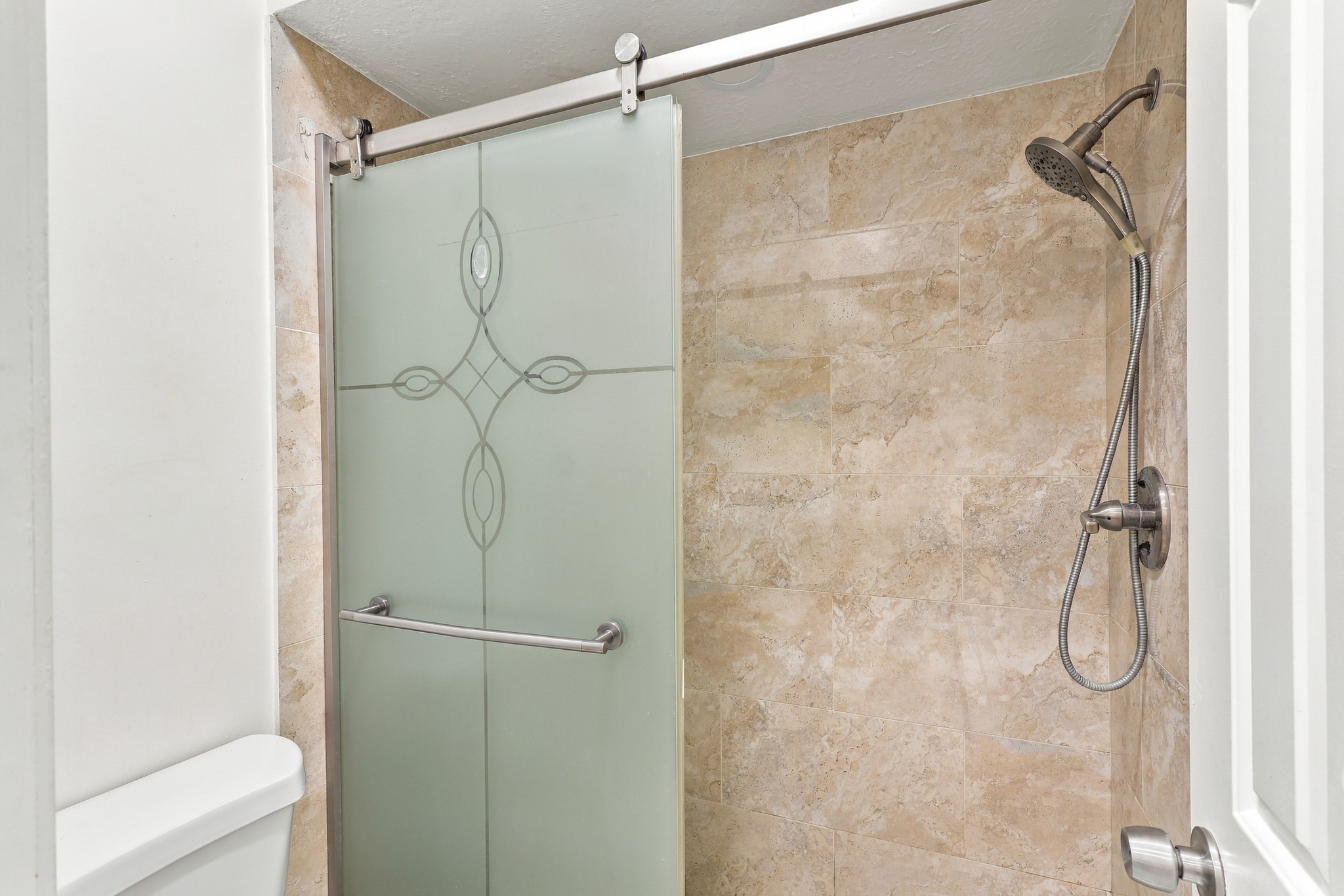 Spacious walk-in shower featuring neutral stone tile extending to the ceiling, a decorative frosted glass sliding door with brushed nickel hardware, and a convenient handheld showerhead.