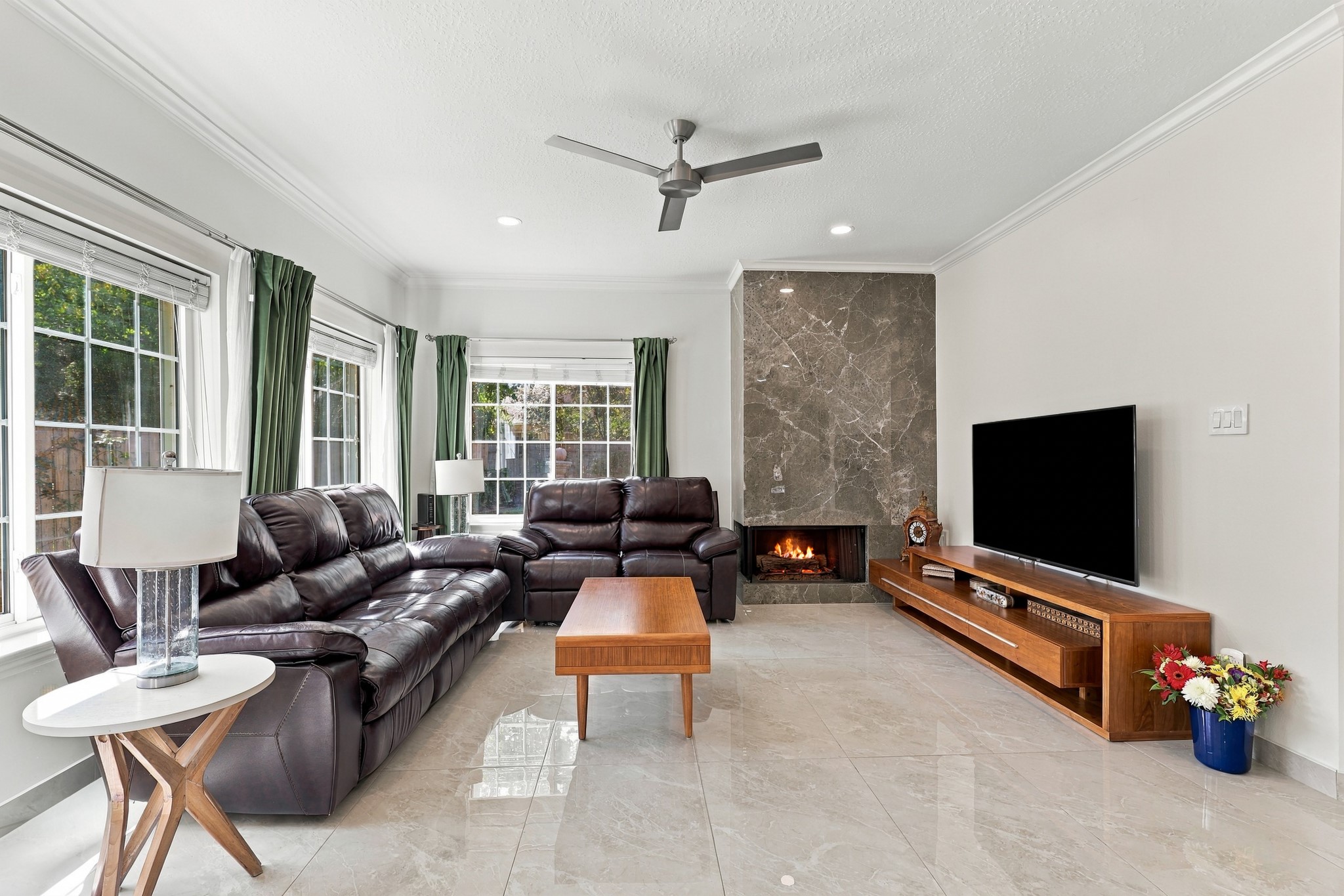 Another view of the inviting living room showcases the generous seating area, abundant natural light, and beautiful marble tile flooring. A floor-to-ceiling fireplace with dramatic surround anchors the space, complemented by crown molding, recessed lighting, and a sleek contemporary ceiling fan. Another view of the inviting living room showcases the generous seating area, abundant natural light, and beautiful marble tile flooring. A floor-to-ceiling fireplace with dramatic surround anchors the space, complemented by crown molding, recessed lighting, and a sleek contemporary ceiling fan.