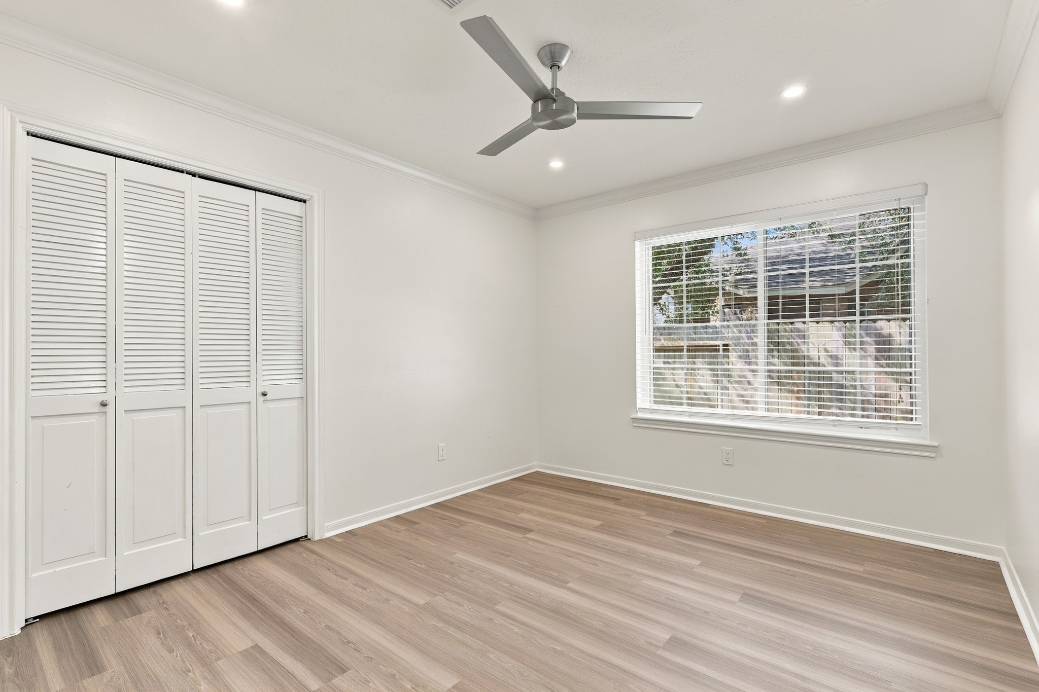 Bright, spacious bedroom featuring wood flooring, crown molding, recessed lighting, and a modern ceiling fan. A large window provides natural light, while a generous double closet with louvered doors offers ample storage.