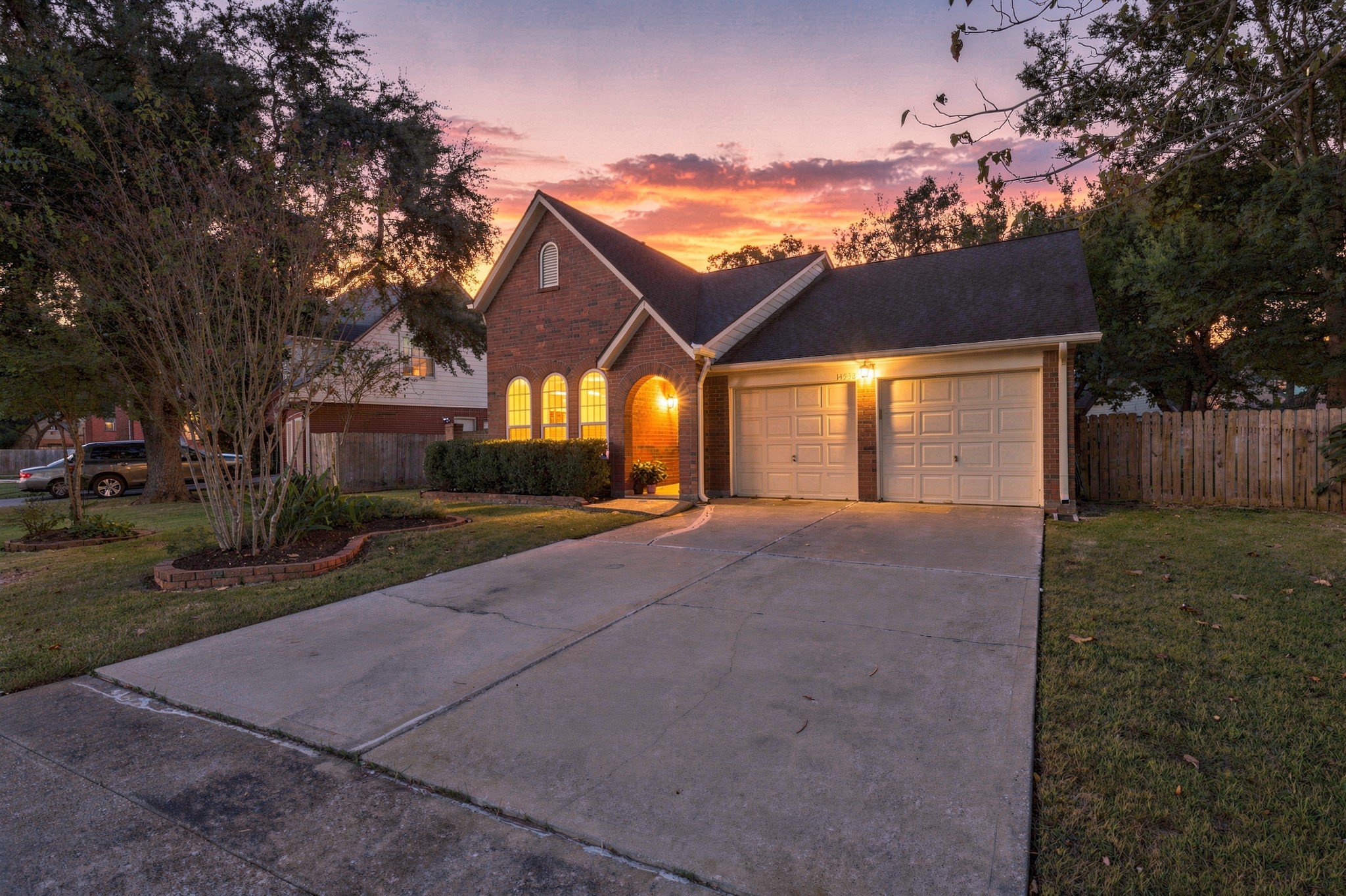 Beautiful view showcasing the home's charming brick exterior, arched entryway, elegant arched windows, and welcoming exterior lighting. Mature trees, landscaped beds, a spacious driveway, and an attached two-car garage complete the inviting curb appeal.