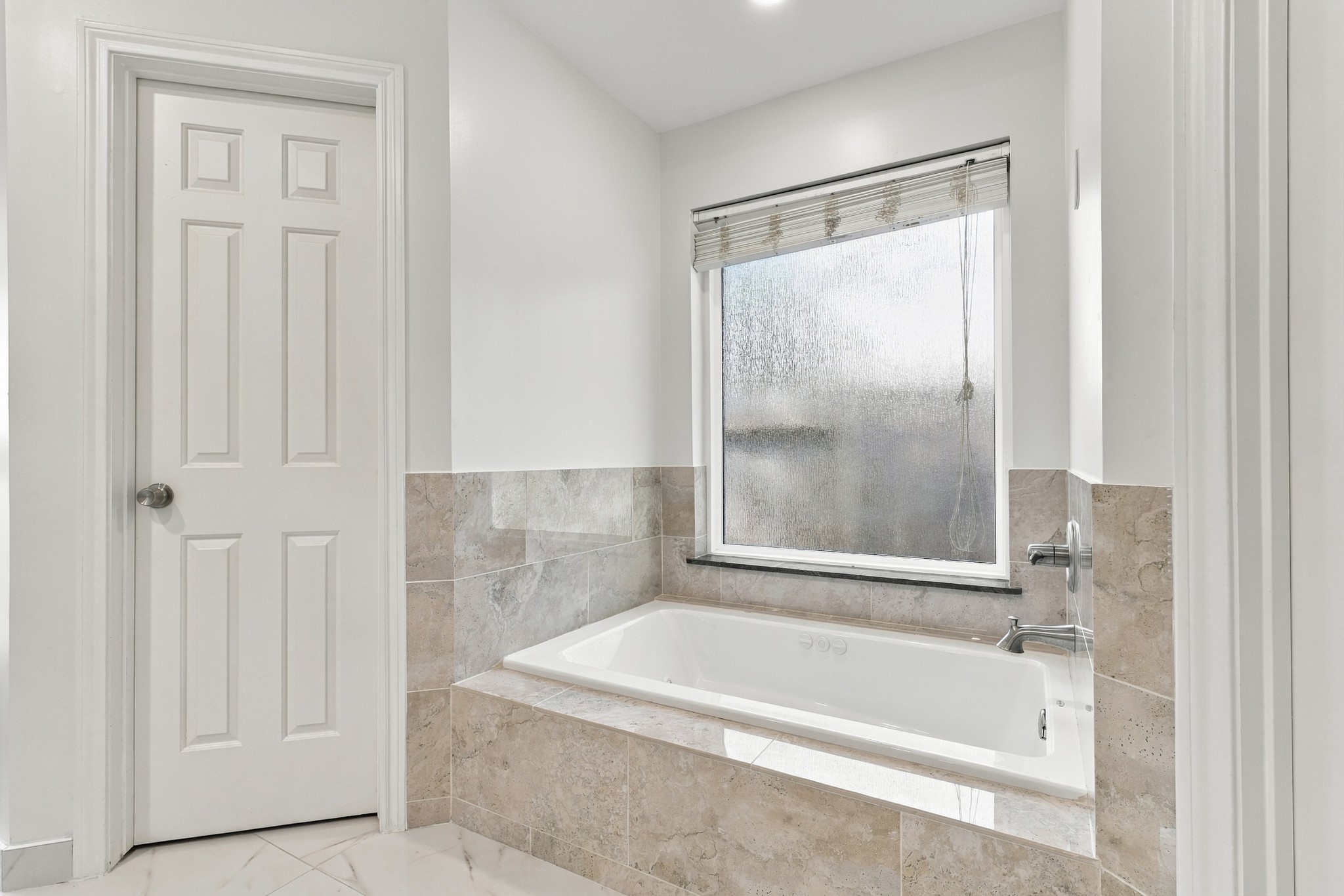 Primary bathroom features a relaxing jetted soaking tub surrounded by neutral stone tile, with a large frosted window providing natural light and privacy. A perfect spot to unwind at the end of the day.