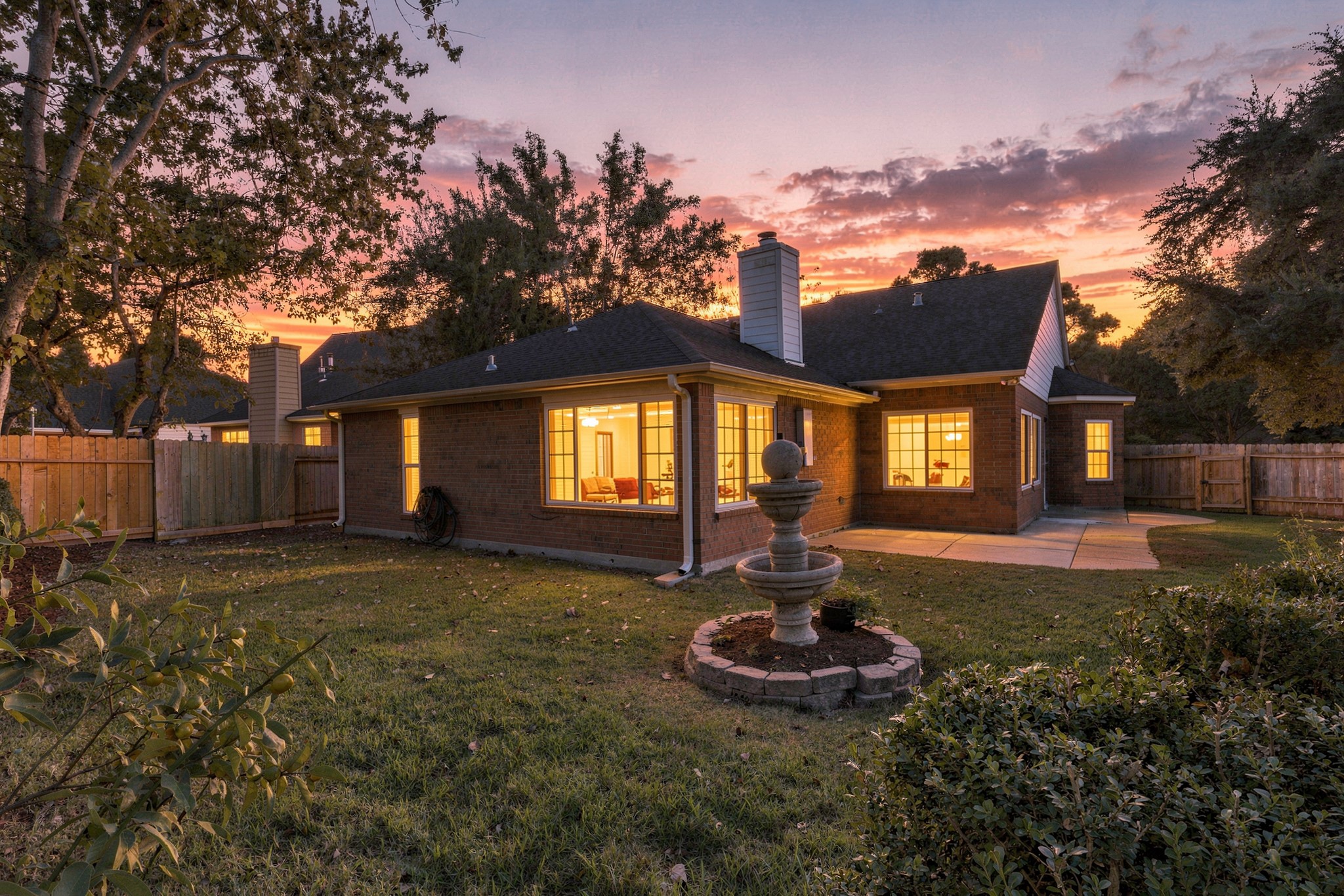 As the sun sets, this beautiful brick home takes on an inviting glow, with warm interior lighting shining through its expansive windows. The backyard features a spacious patio, mature trees, a decorative tiered fountain, and a fully fenced lawn. A charming setting for relaxing evenings, outdoor gatherings, and enjoying the comforts of home.