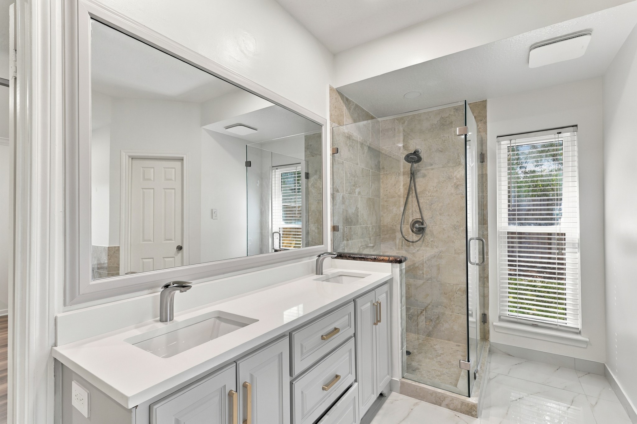 The primary bathroom features a spacious double-sink vanity with sleek countertops, gray cabinetry, gold-toned hardware, and an oversized mirror. A separate glass-enclosed walk-in shower with neutral stone tile completes the bright, modern space.