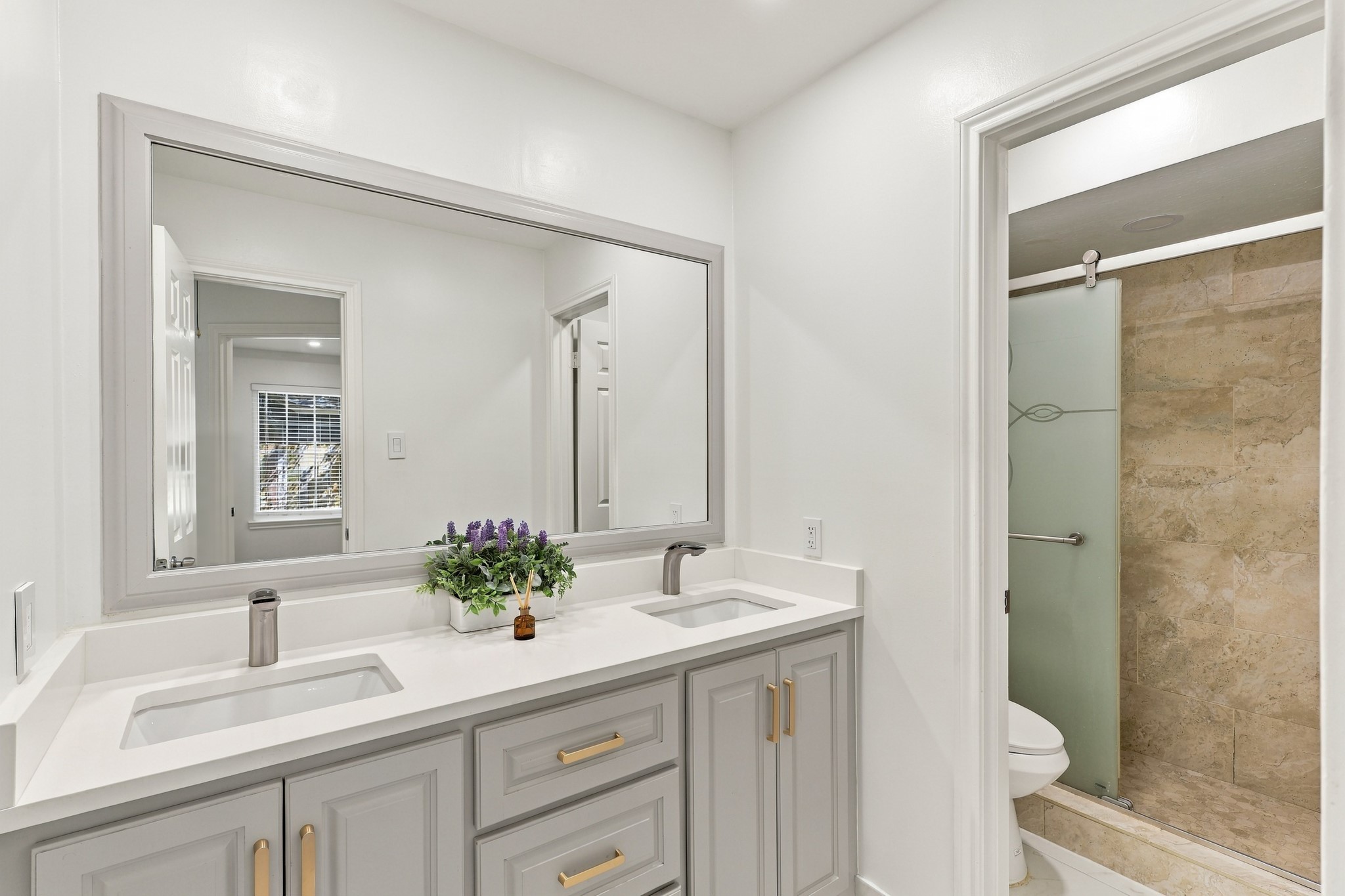 Beautifully updated bathroom featuring a spacious double-sink vanity with sleek countertops, gray cabinetry, brushed gold hardware, and a large framed mirror. The adjoining walk-in shower features floor-to-ceiling tile and a frosted glass enclosure.