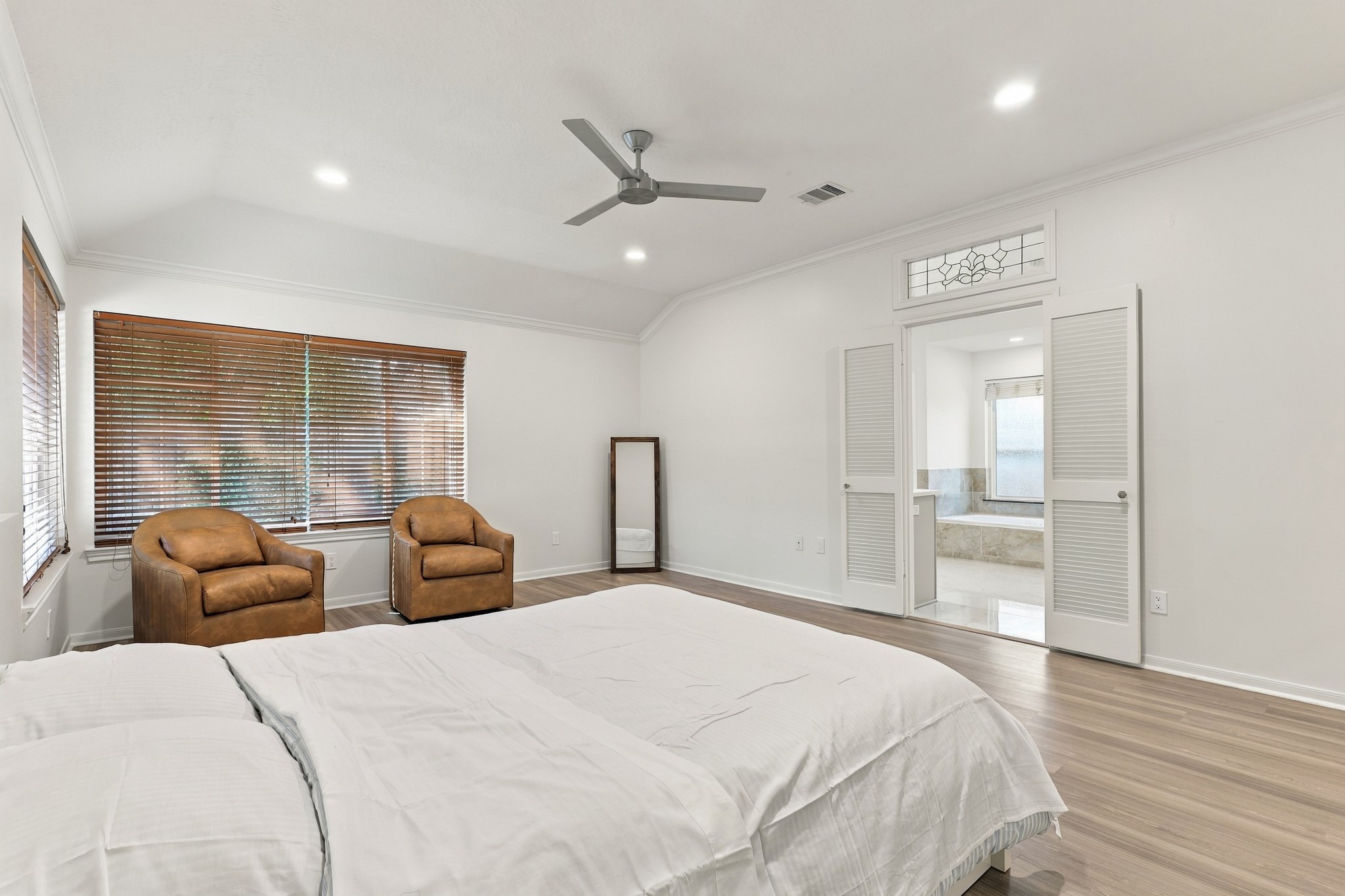Another view of the spacious primary suite highlights the vaulted ceiling, crown molding, wood flooring, and comfortable sitting area. Decorative glass transom and double louvered doors create an elegant entrance to the adjoining primary bathroom.