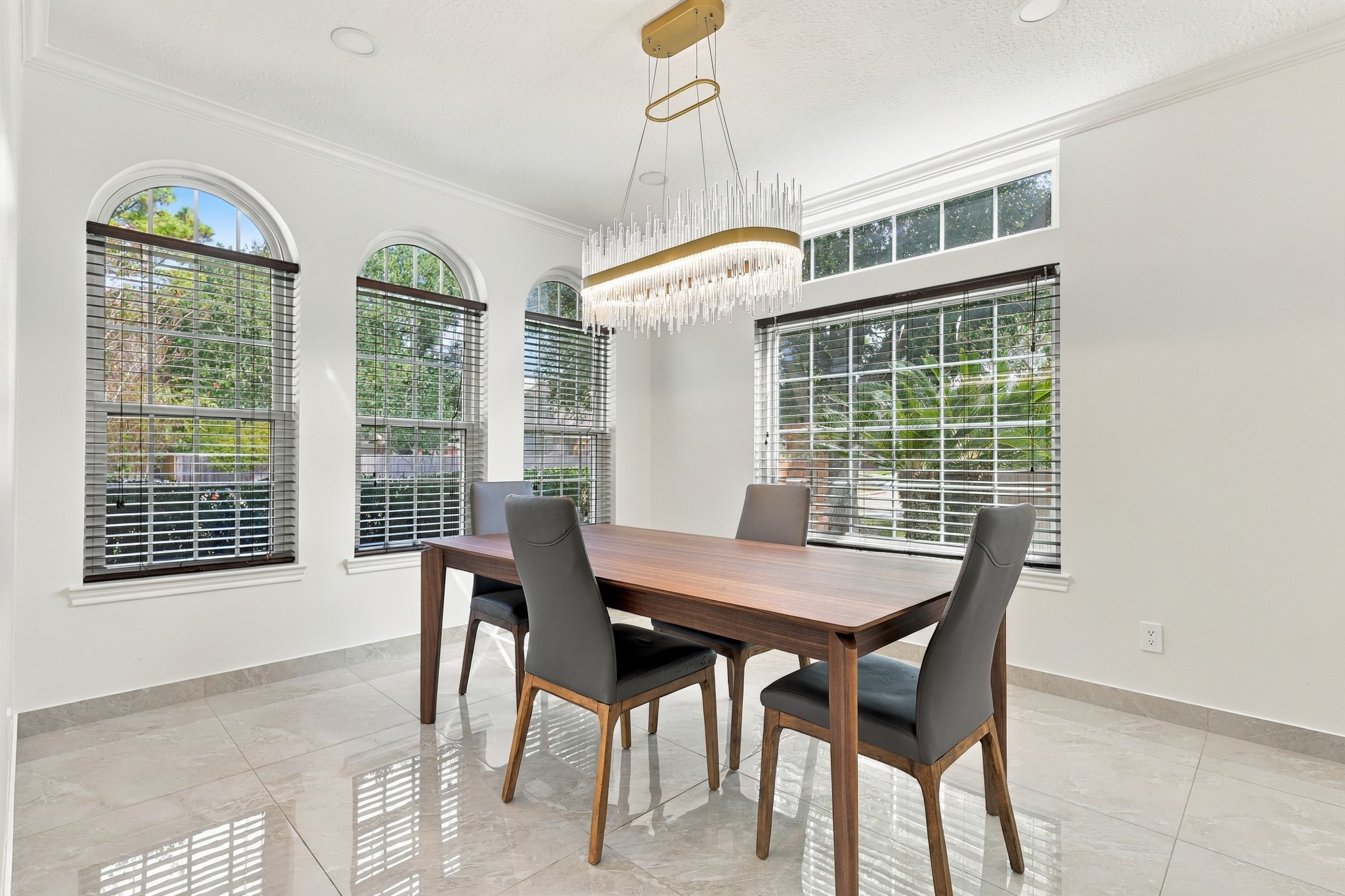 Elegant formal dining room filled with natural light from three arched windows and an oversized picture window with transom detailing. A stunning crystal-accented chandelier, crown molding, recessed lighting, and glossy marble tile flooring create a sophisticated setting for dining and entertaining. Elegant formal dining room filled with natural light from three arched windows and an oversized picture window with transom detailing. A stunning crystal-accented chandelier, crown molding, recessed lighting, and glossy marble tile flooring create a sophisticated setting for dining and entertaining.