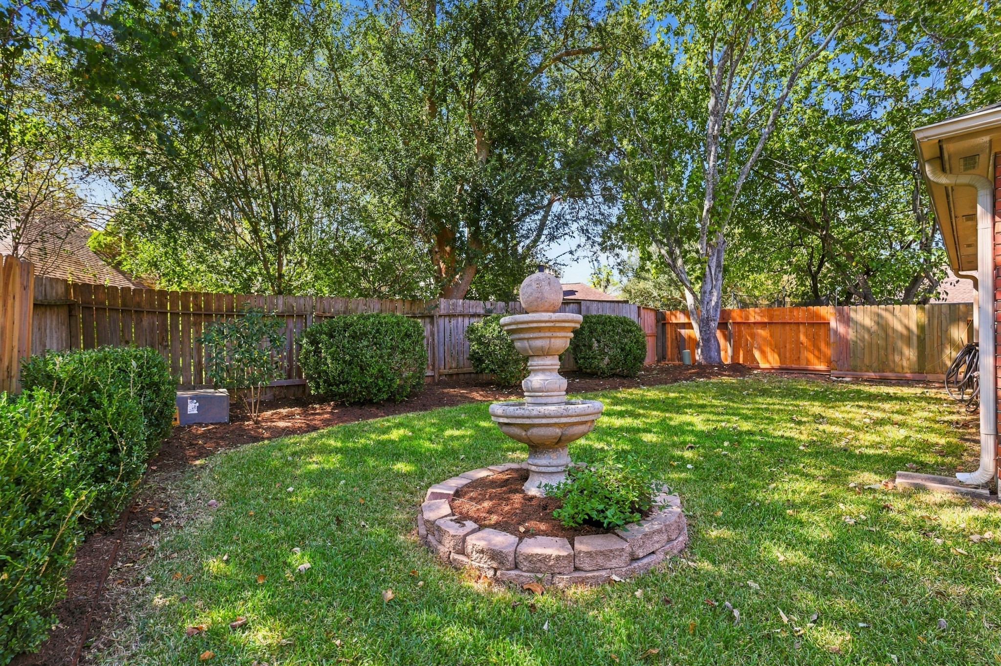 The backyard offers a peaceful, private-feeling retreat surrounded by mature shade trees, established landscaping, and wood fencing. A decorative tiered fountain serves as the centerpiece of the lawn, complemented by neatly maintained shrubs and plenty of green space for outdoor enjoyment.
