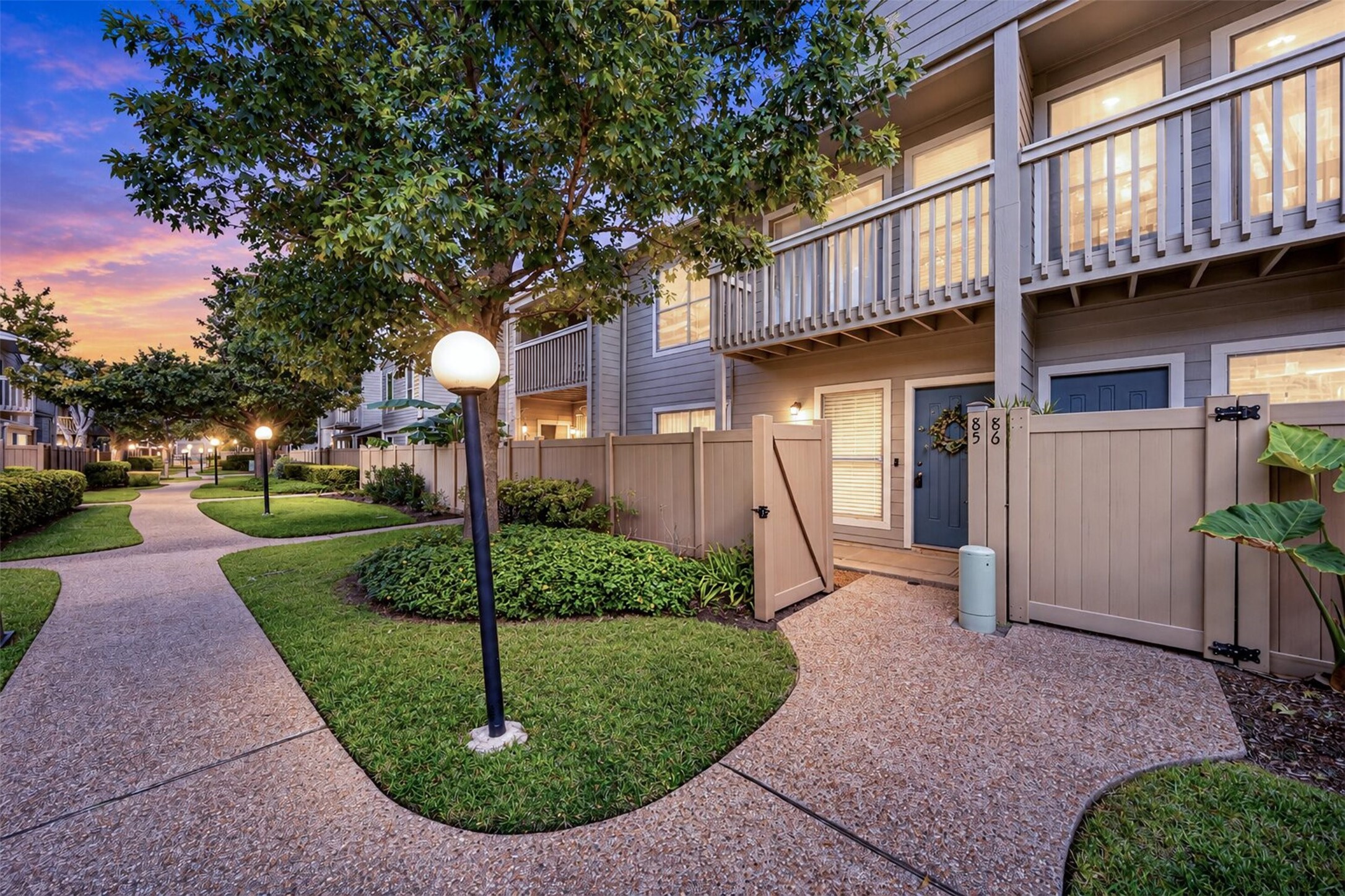 Twilight settles over Unit 85 as warm light glows from the windows and balcony. The gated entry, tree-lined walkways, and lampposts give the community an evening glow.