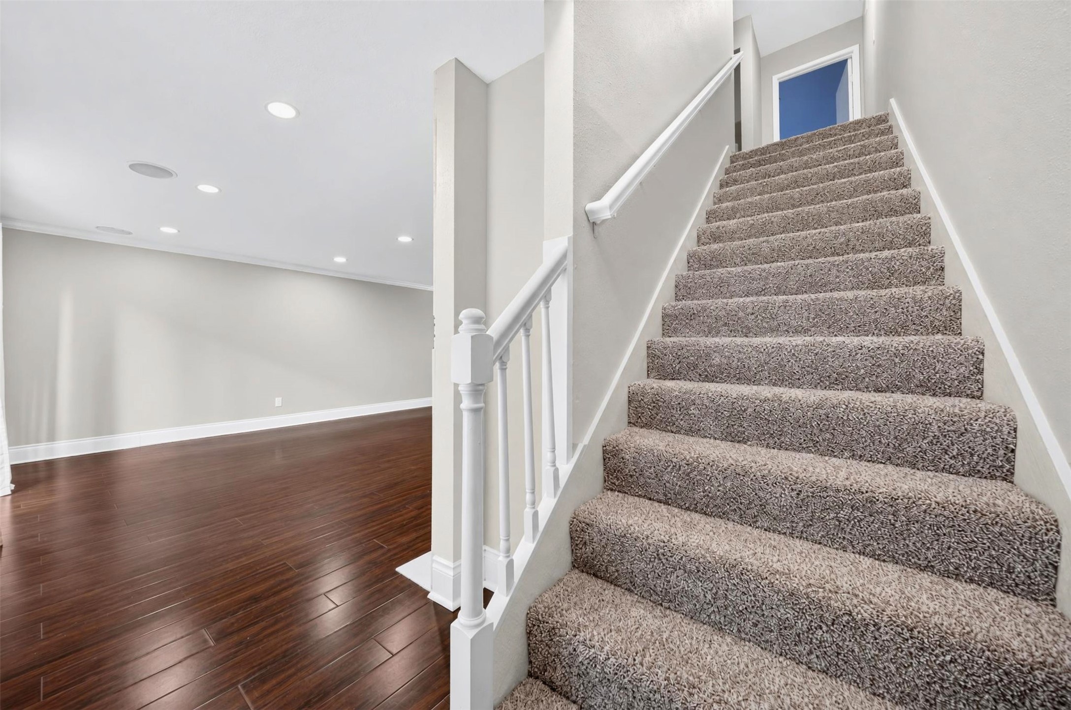 Carpeted stairs with a white wood railing climb from the main level, where dark hardwoods and recessed lights continue into the living area.