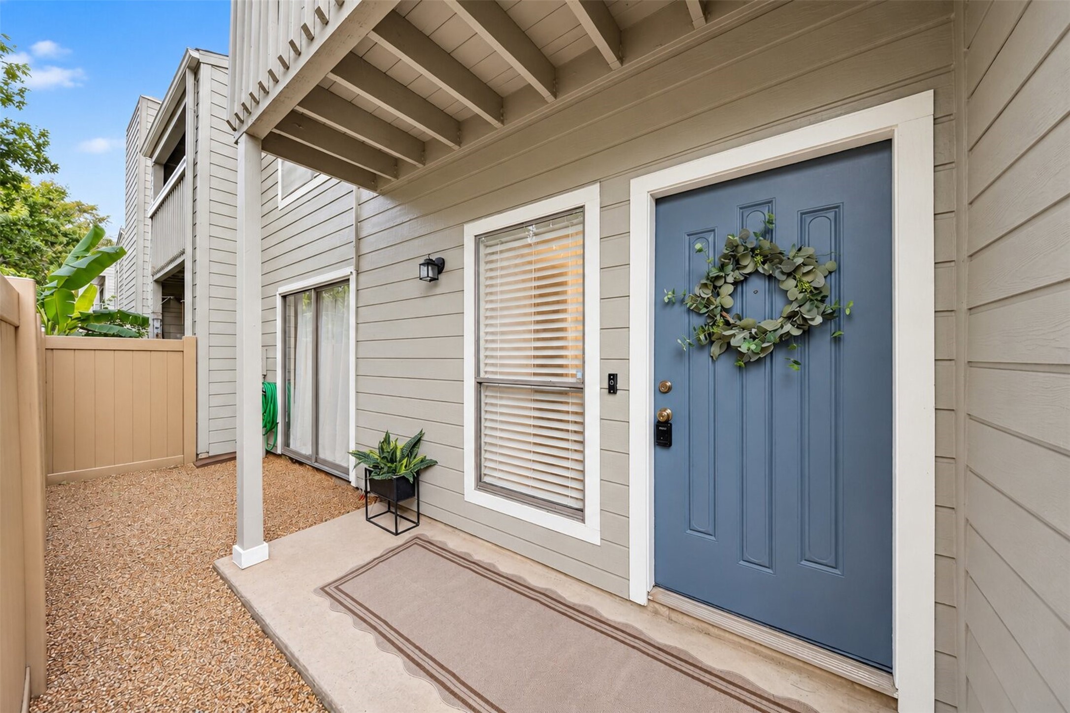 A covered front porch leads to the blue entry door, dressed with a eucalyptus wreath and keypad lock. Beside it, a private fenced side patio with sliding glass door extends the living space outdoors.