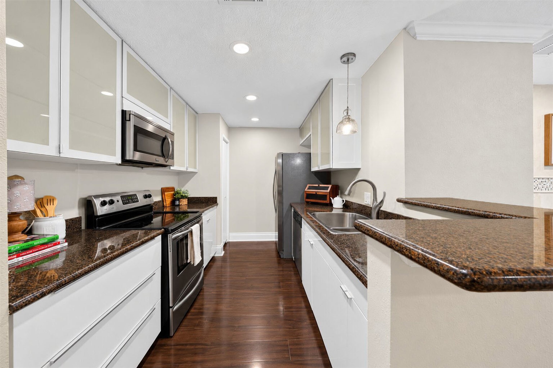 The galley kitchen pairs glossy white cabinetry with dark granite counters and stainless appliances  smooth-top range, built-in microwave, and French-door fridge. A double sink and raised breakfast bar open to the dining room.