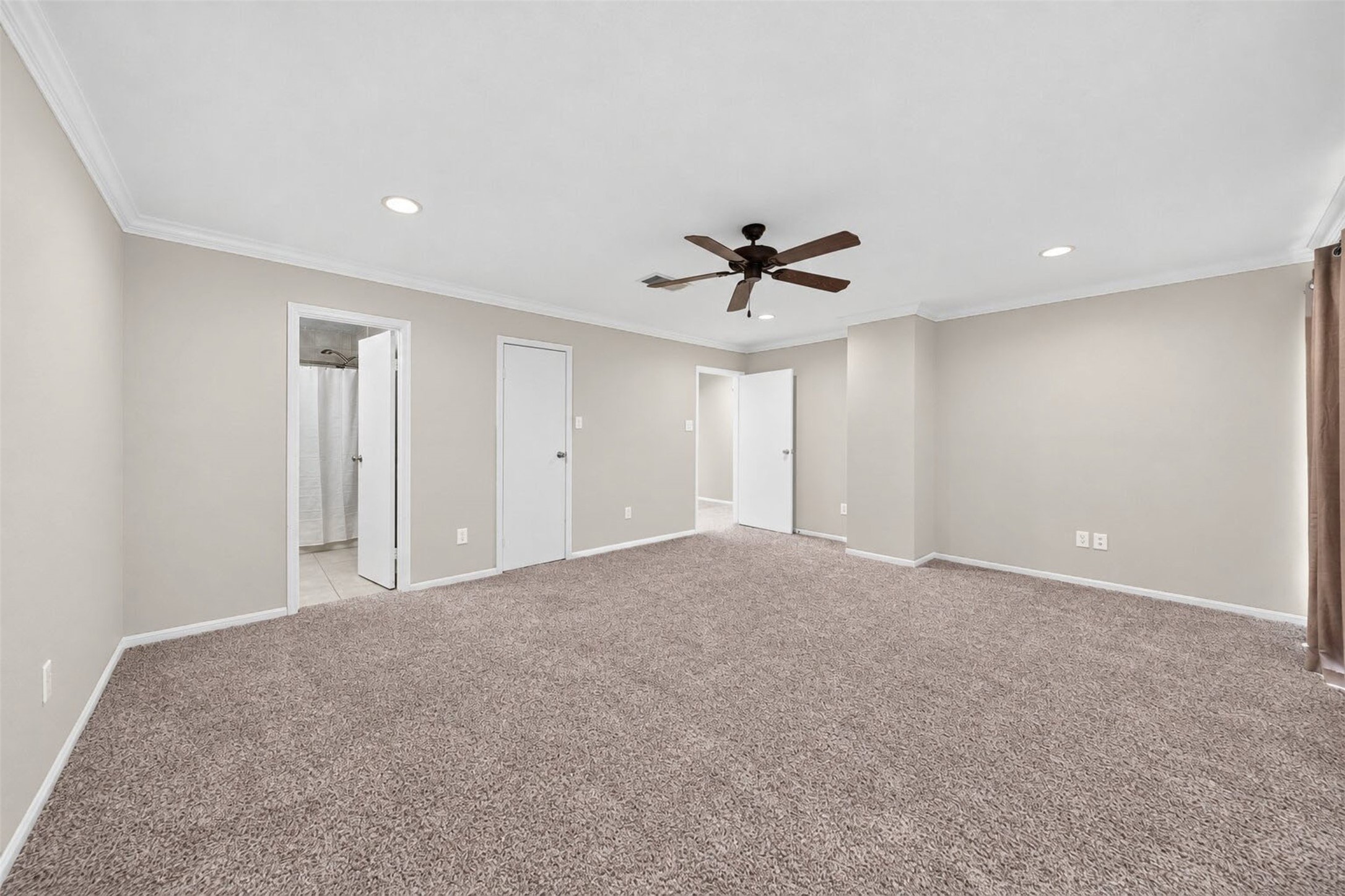 From the window corner, the primary bedroom shows its ensuite bath doorway, a closet door, and the hall entry. Plush carpet and a ceiling fan throughout.