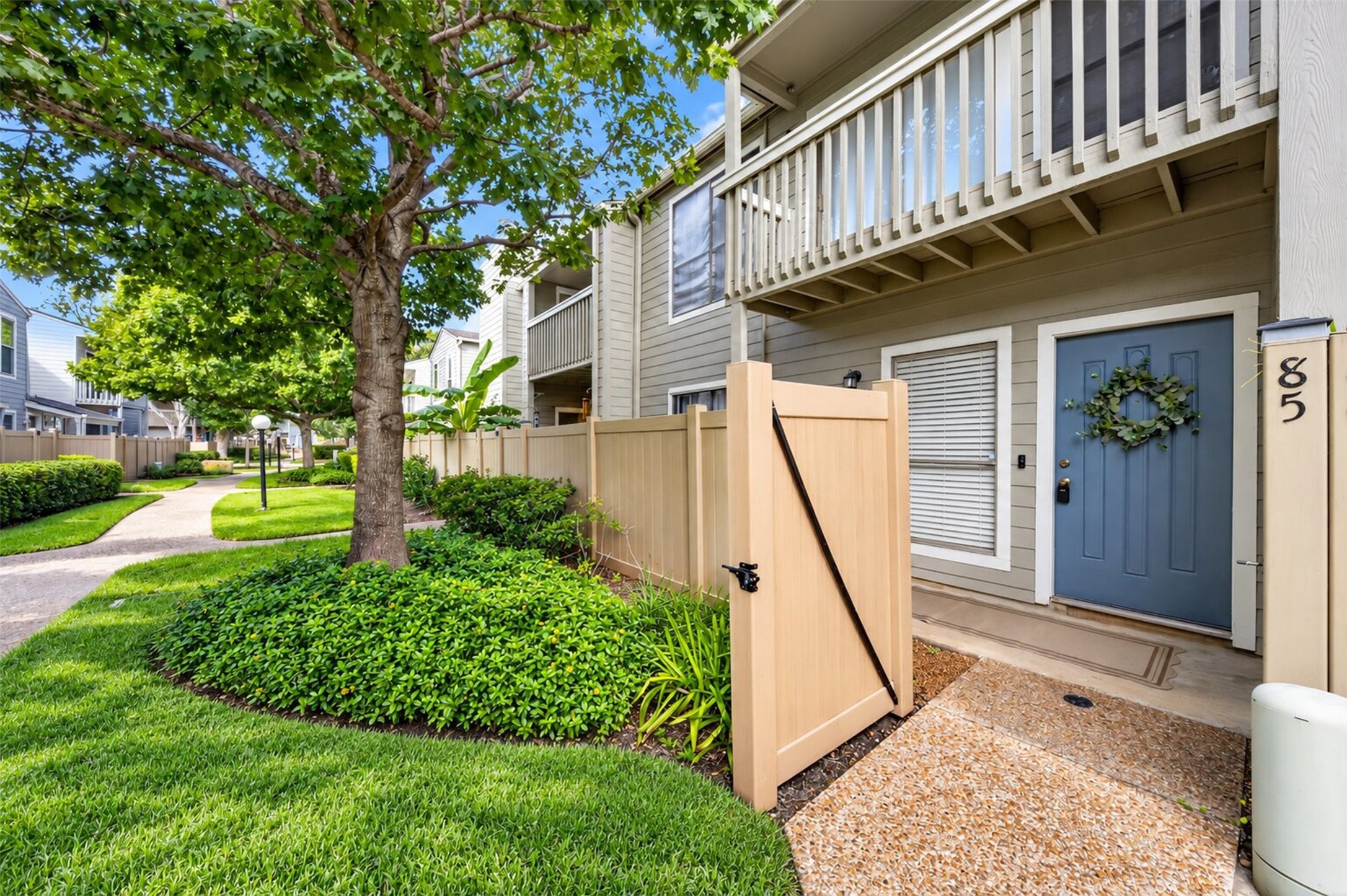 Welcome home to Unit 85: a slate-blue front door with a greenery wreath, a gated front patio, and a second-story balcony, all shaded by mature trees in a manicured, walkable community.