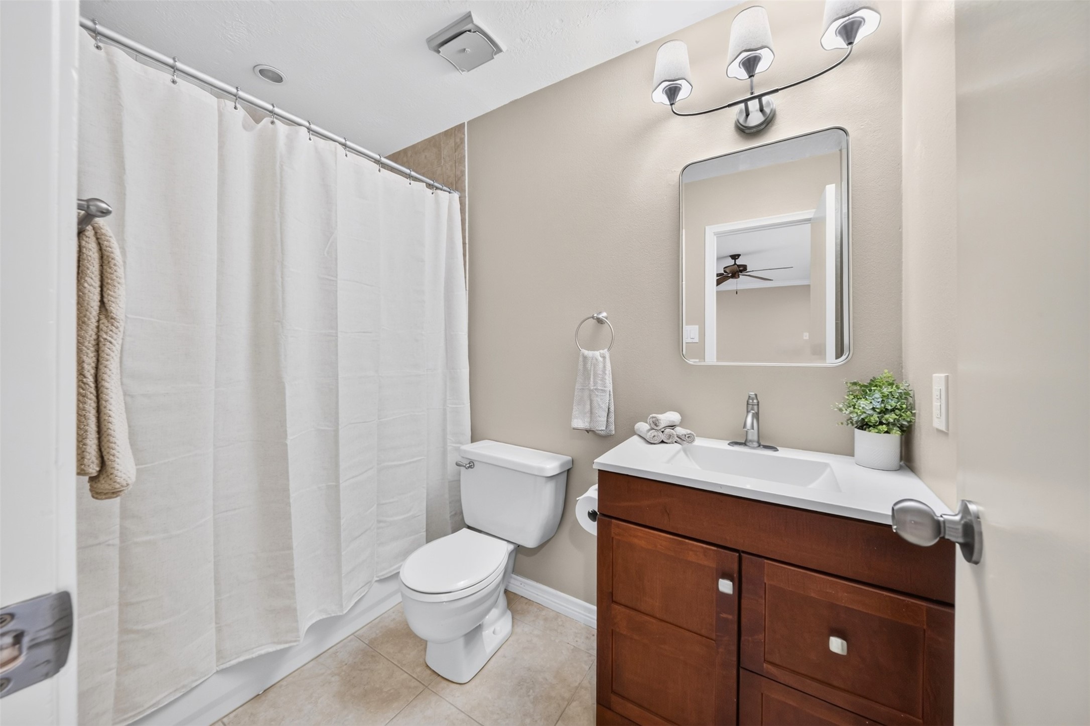 This full bath pairs a rich dark-wood vanity with a white countertop sink, a rounded mirror, and a tub/shower with a linen curtain.