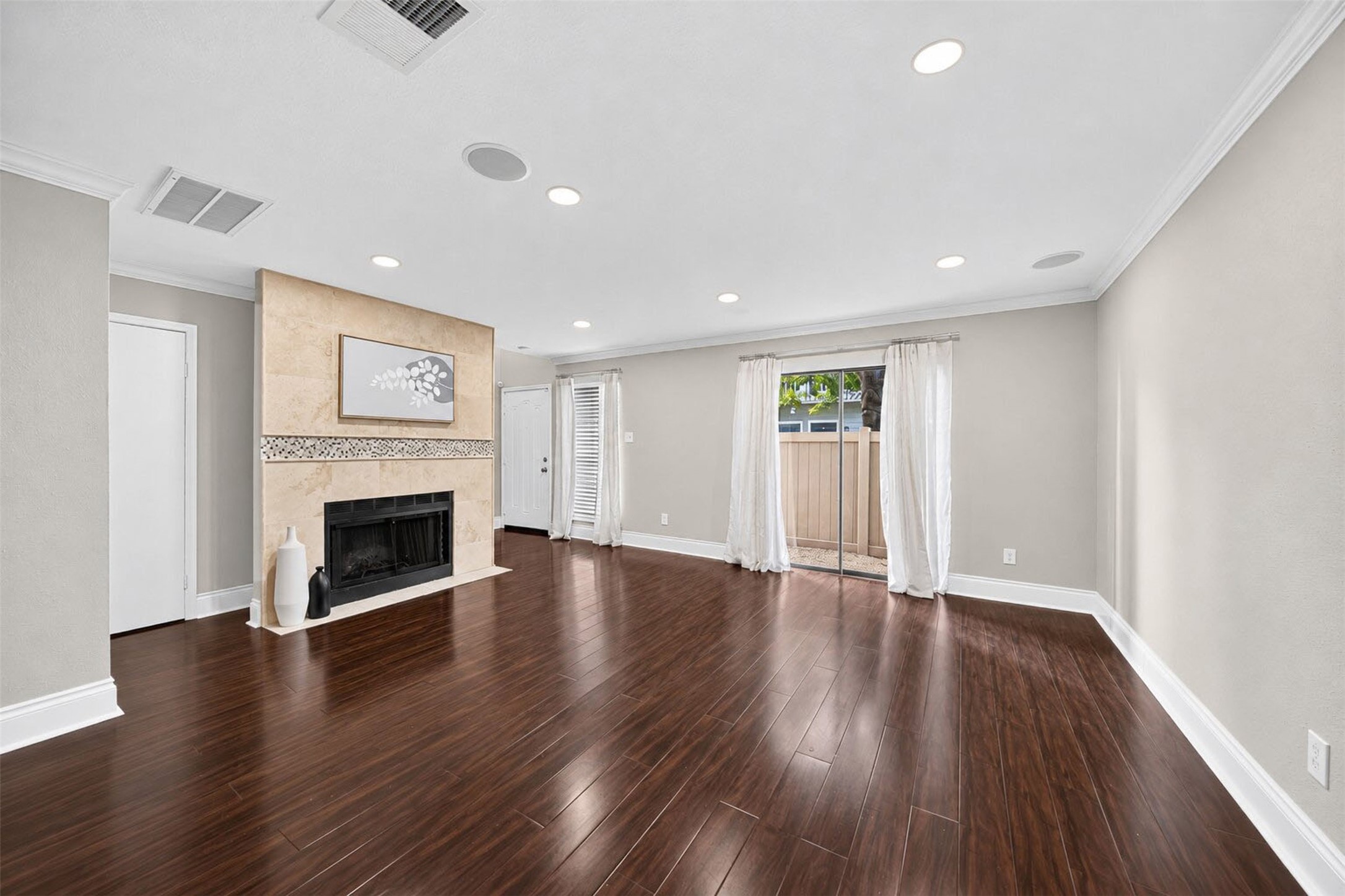 From the dining side, the living room centers on the tiled fireplace, with the front door beyond and a sliding glass door opening to the side patio. Dark wood floors and in-ceiling speakers run throughout.