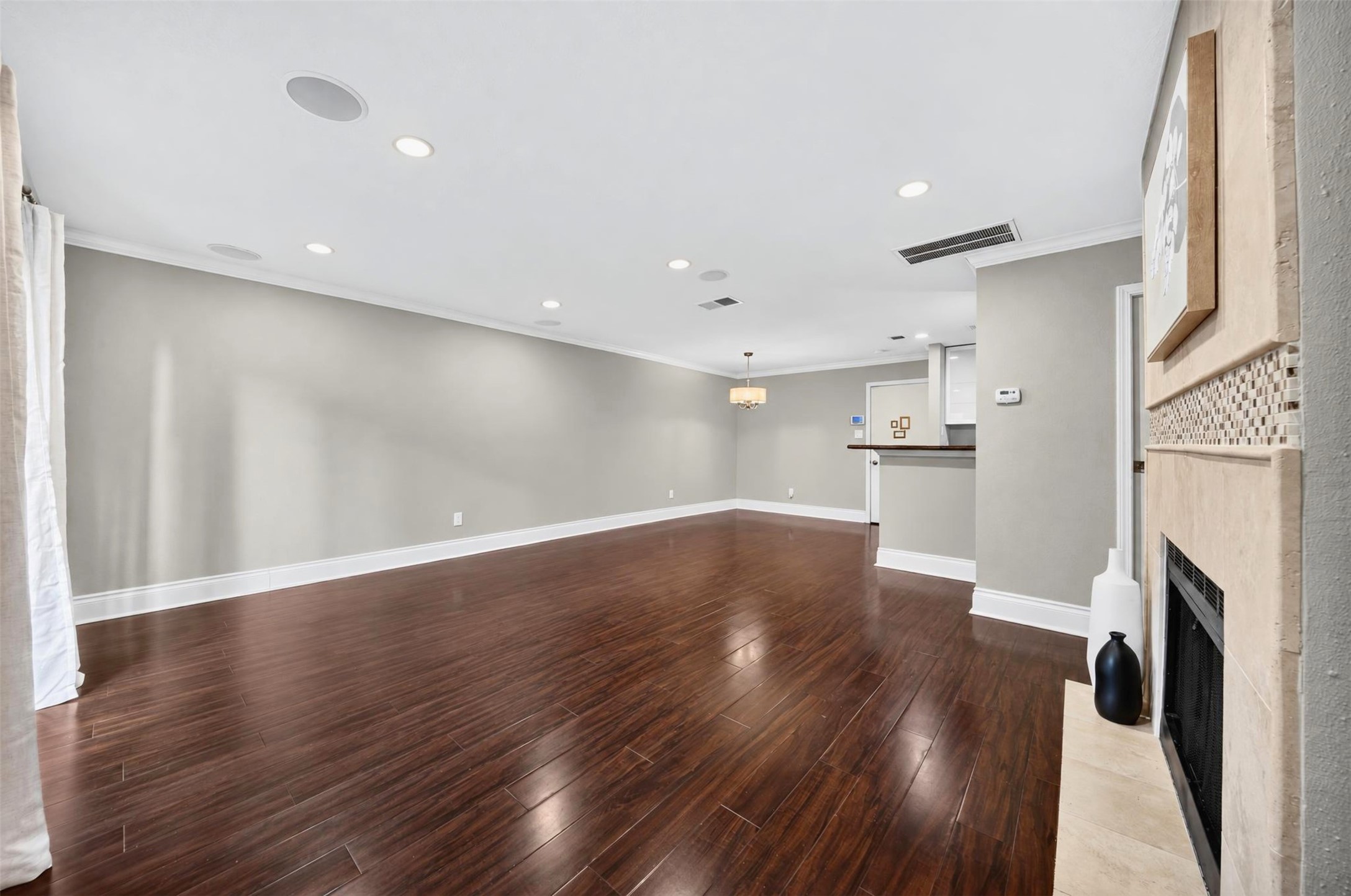 Looking down the length of the open living space toward the dining area and kitchen bar, with the fireplace hearth at right. Gleaming dark hardwoods and recessed lighting throughout.