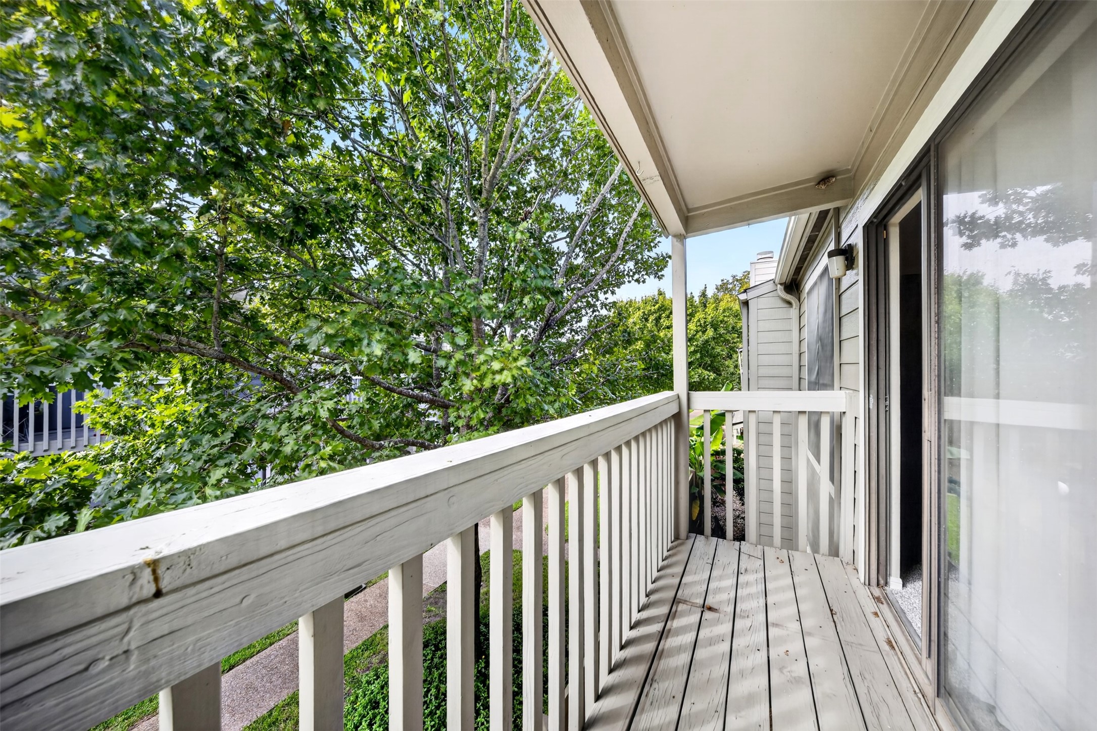 A second covered balcony off the front of the unit, with a wood deck and railing facing mature trees and the community walkway.