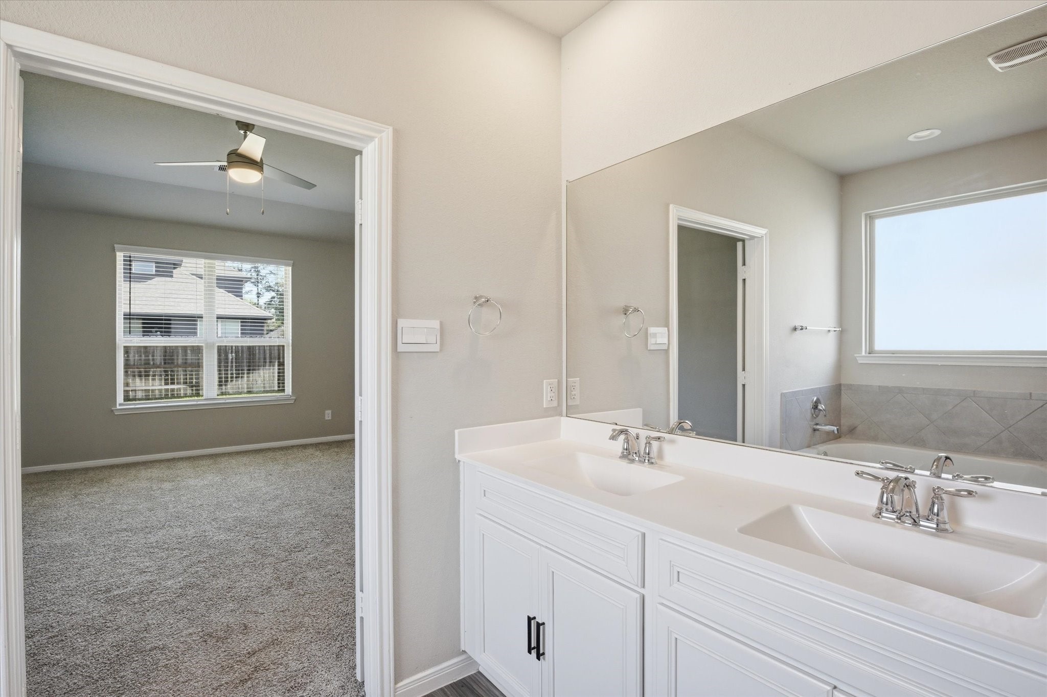 This photo shows a bright bathroom with dual sinks, a large mirror, and a window providing natural light. It connects to a carpeted bedroom featuring a ceiling fan and a window with a view of the outdoors.