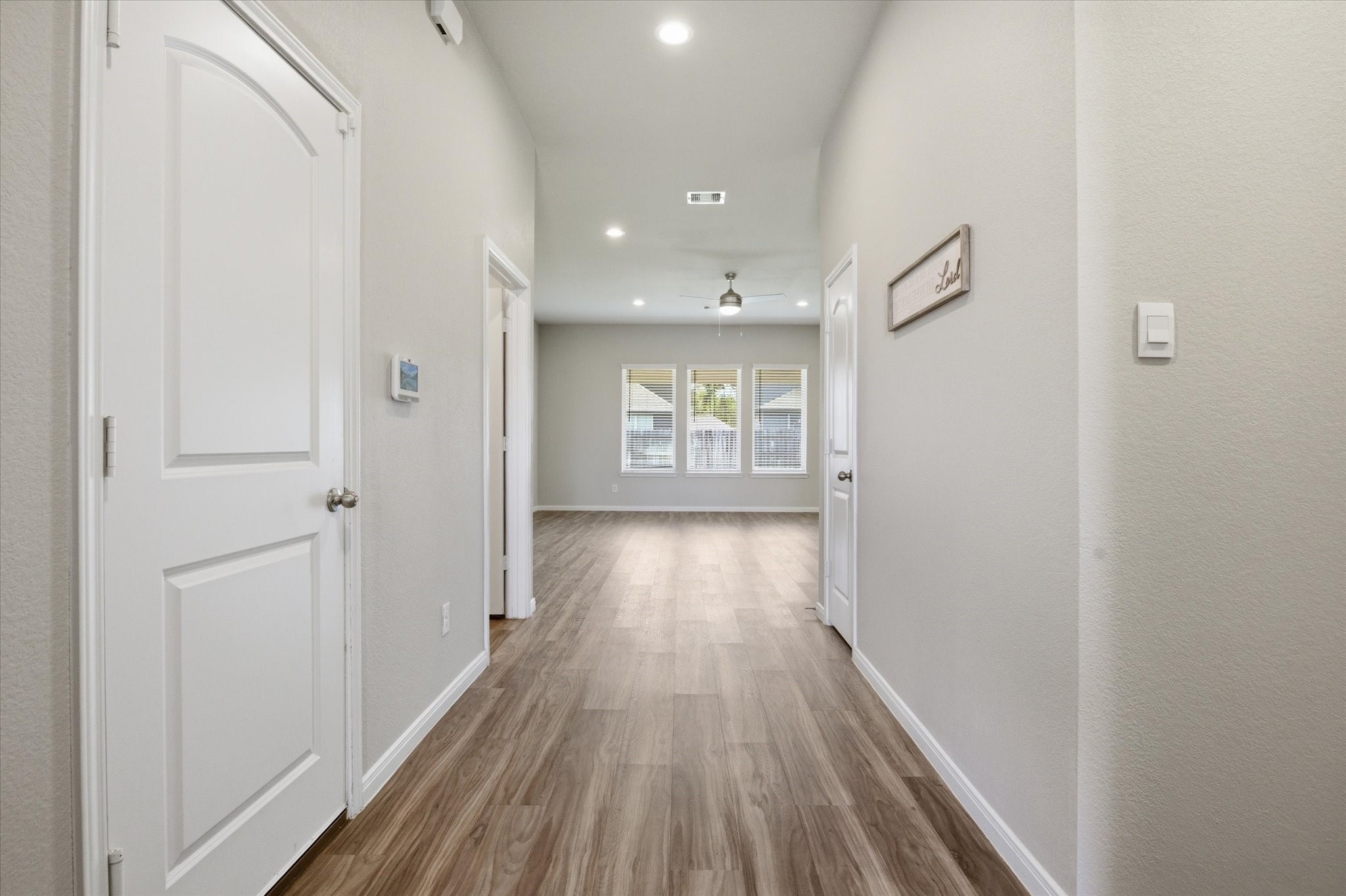 This photo showcases a spacious open living area with dark wood flooring, neutral walls, and ample natural light from multiple windows. The room features a ceiling fan and recessed lighting, with an open view into a modern kitchen area with a large counter and sink.