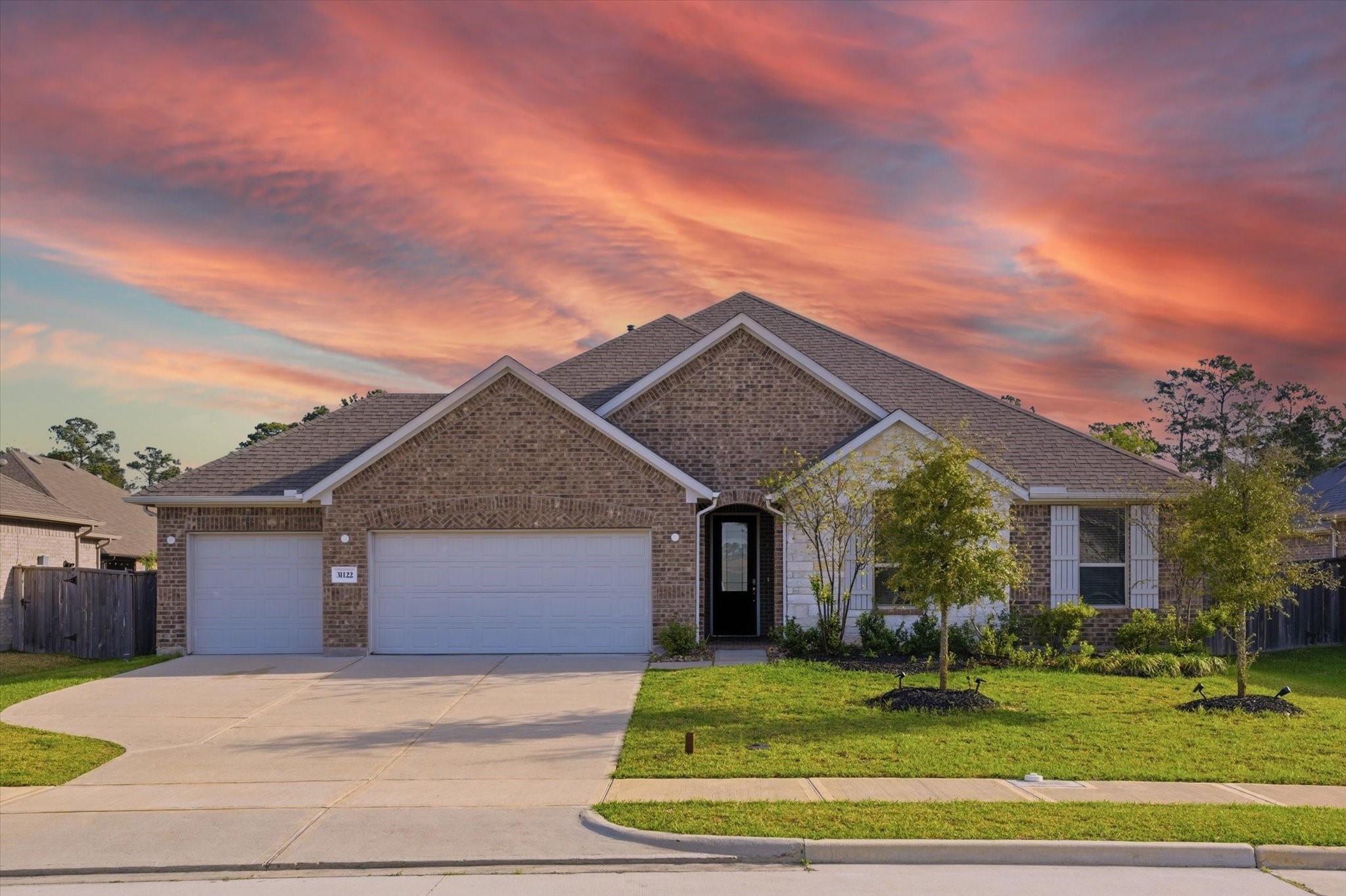 This is a modern, single-story brick home with a spacious driveway leading to a two-car garage. The exterior features well-maintained landscaping and warm, inviting lighting, set against a picturesque sunset sky.