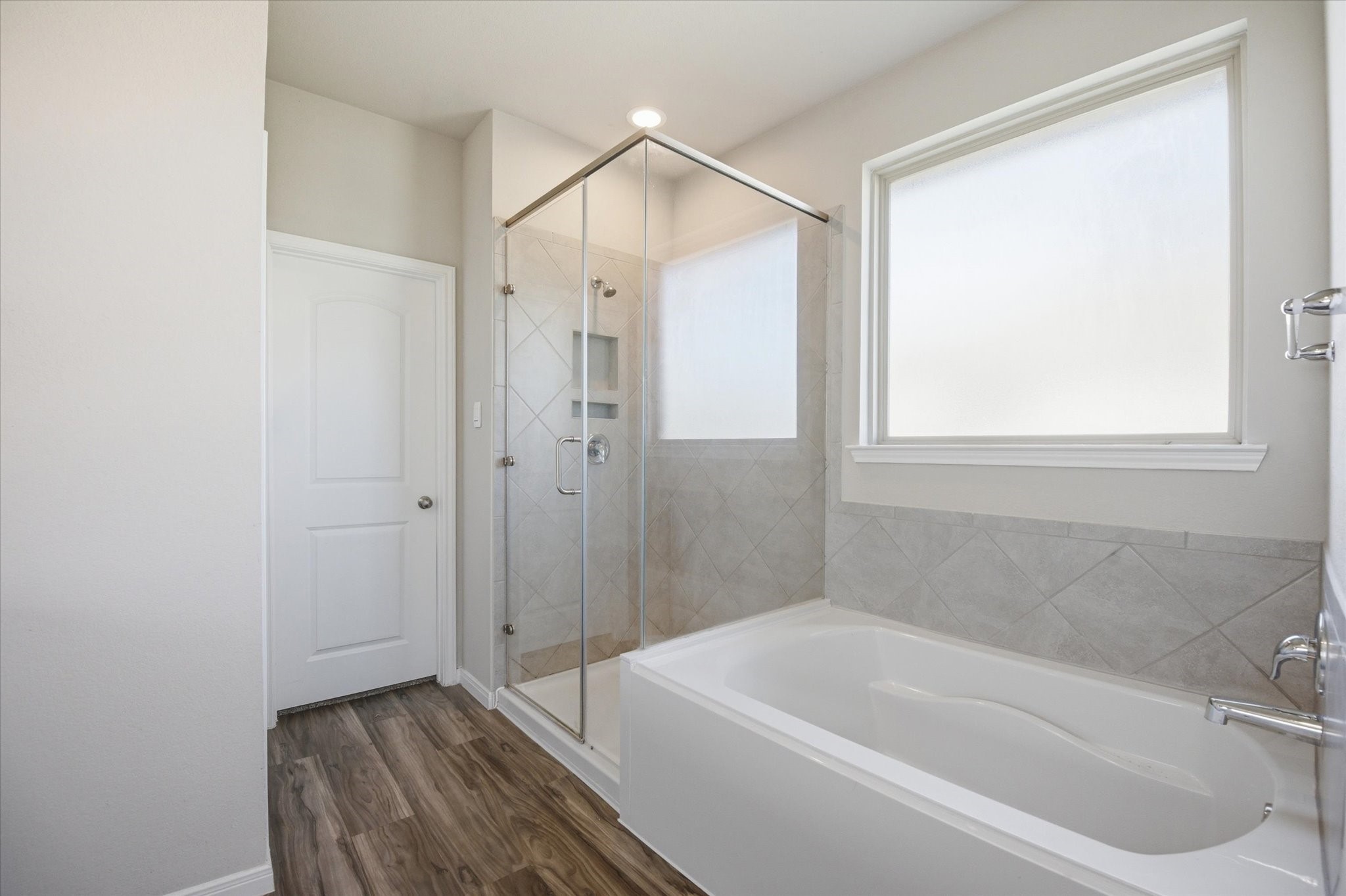 This bathroom features a modern design with a glass-enclosed shower, a large tub, and sleek tile work. Natural light fills the space through a frosted window, creating a bright and inviting atmosphere. The wood-look flooring adds warmth and elegance.