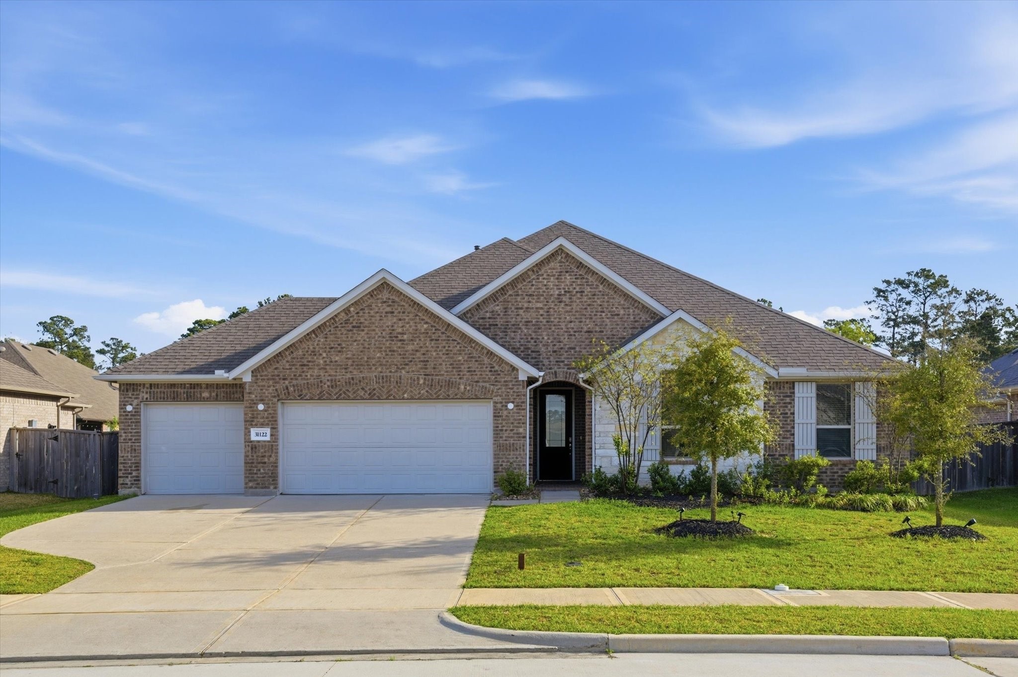 This is a modern, single-story brick home with a pitched roof, featuring a three-car garage and a well-maintained front yard with young trees and shrubs. It's located in a suburban neighborhood with similar homes.