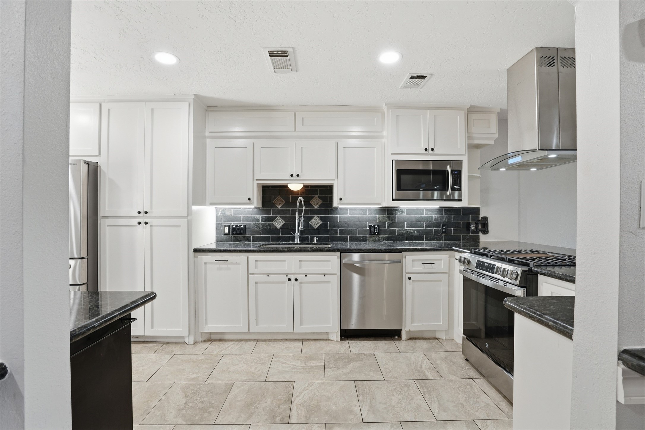 Modern kitchen with white cabinetry, stainless steel appliances, sleek black tile backsplash, and under cabinet lighting. Features ample storage, a built in microwave, and a large gas range.