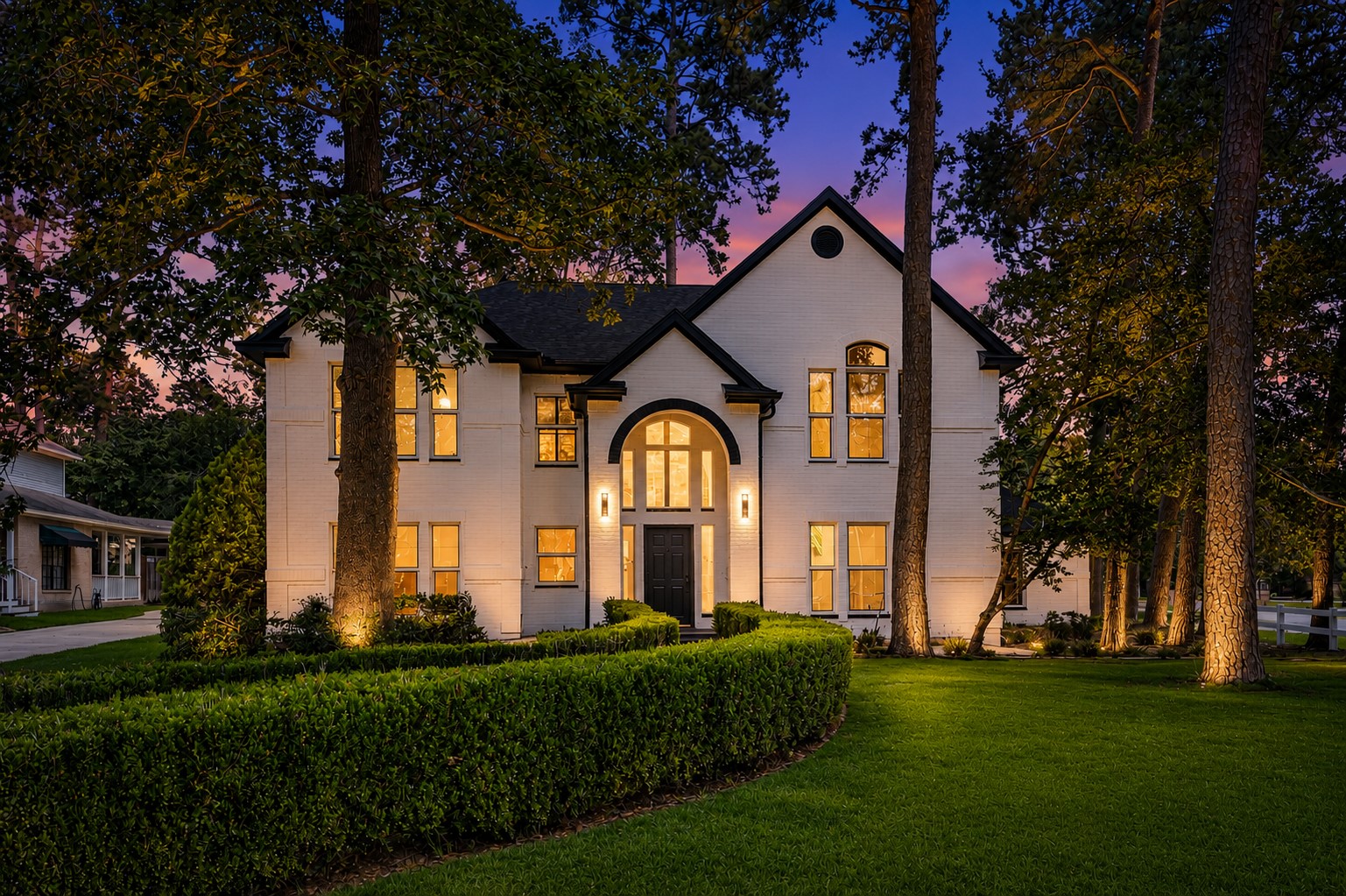 Elegant two-story home with a striking white brick exterior, nestled among mature trees and lush greenery, featuring arched windows and a welcoming entryway. *Photo Digitally enhanced.