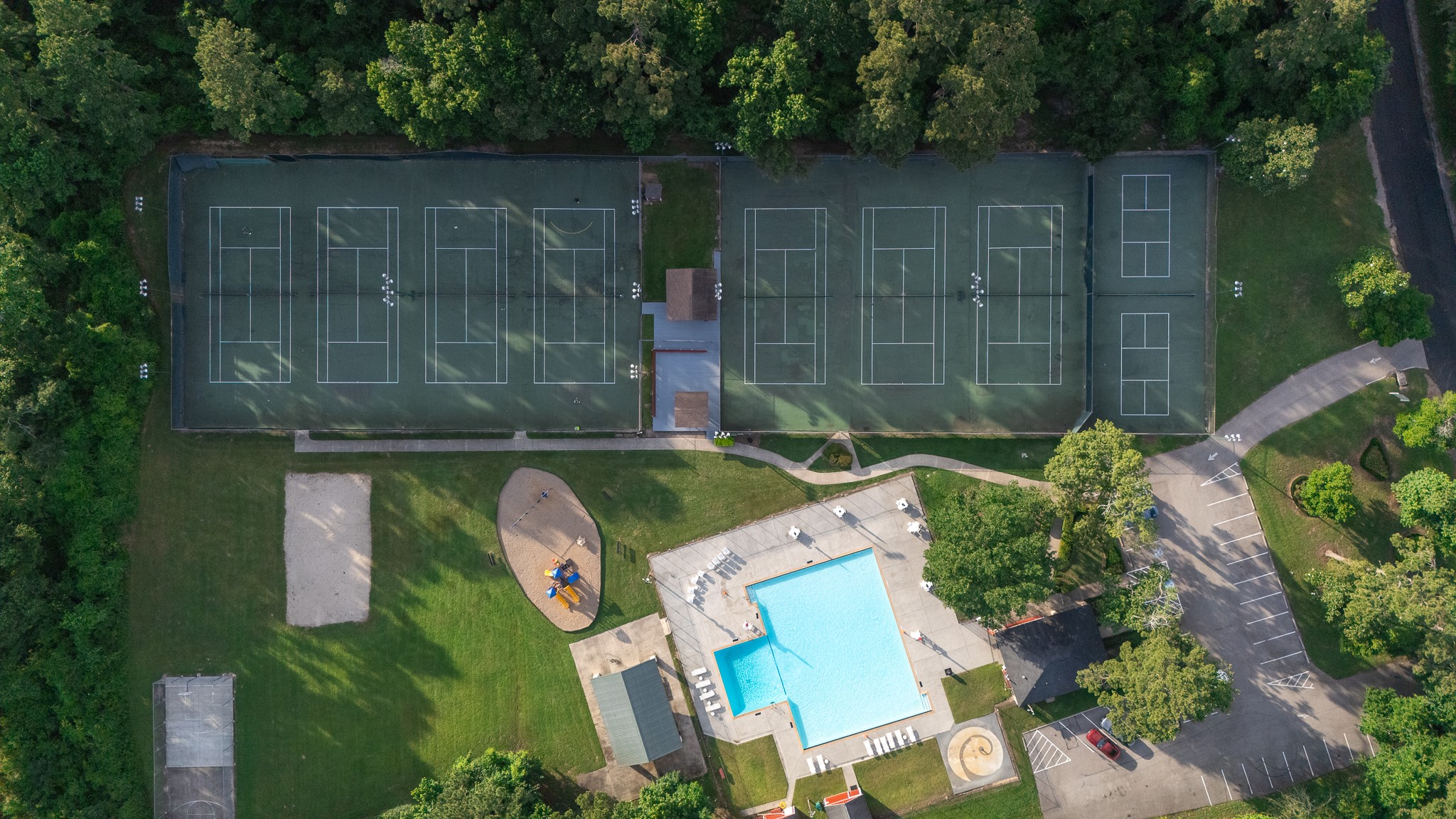 This aerial view showcases a vibrant community recreation area featuring multiple tennis courts, a large swimming pool, a playground, and ample parking, all surrounded by lush greenery.