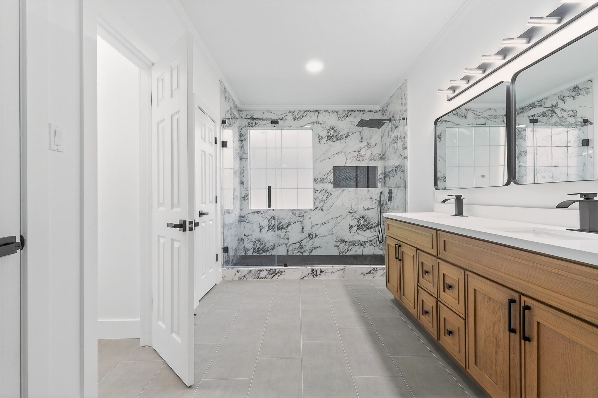 Luxurious bathroom featuring a spacious walk-in shower with elegant marble walls, dual vanity with sleek black fixtures, and ample storage in warm wood cabinetry.