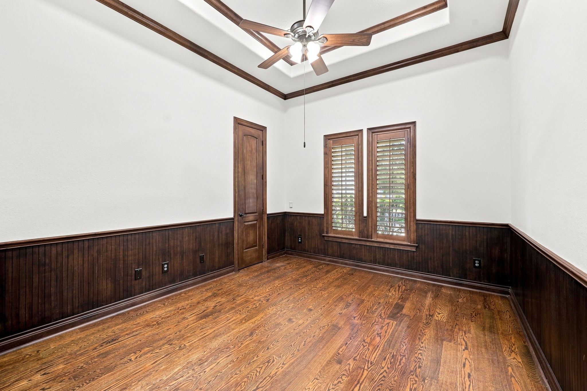 The spacious home office is beautifully appointed with rich wood flooring, a dramatic raised ceiling accented by detailed crown molding, and elegant wainscoting. Plantation shutters frame the windows, adding timeless sophistication while providing privacy and natural light. A generously sized storage closet completes this exceptional and highly functional workspace.