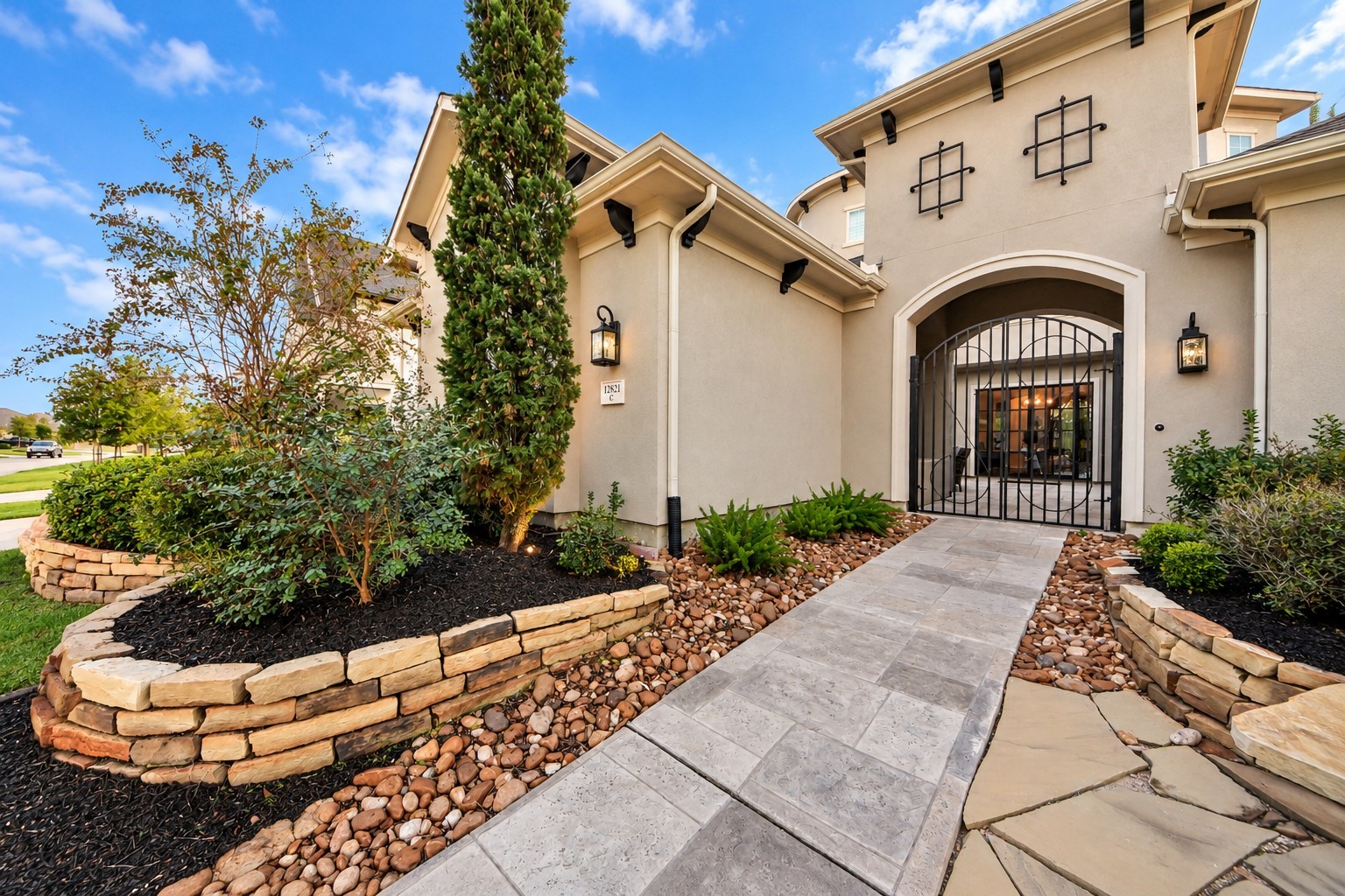 Inviting courtyard entrance featuring elegant double wrought-iron gates, Mediterranean-inspired stucco architecture, lush landscaping, natural stone accents, and decorative rock beds.