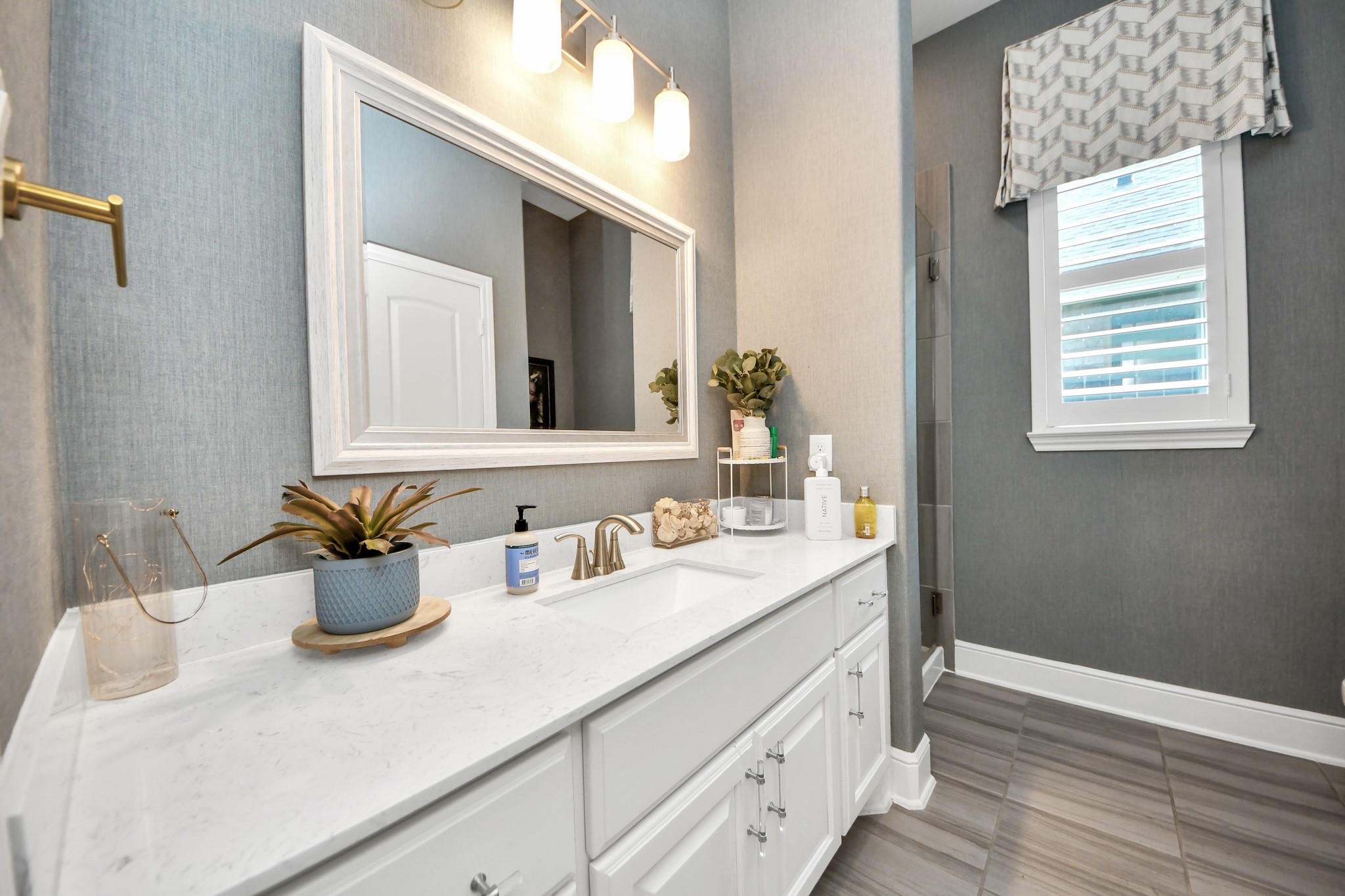 Third full bathroom on the first floor, featuring quartz countertops, decorative wallpaper, plantation shutters, and a walk-in shower with frameless glass enclosure.