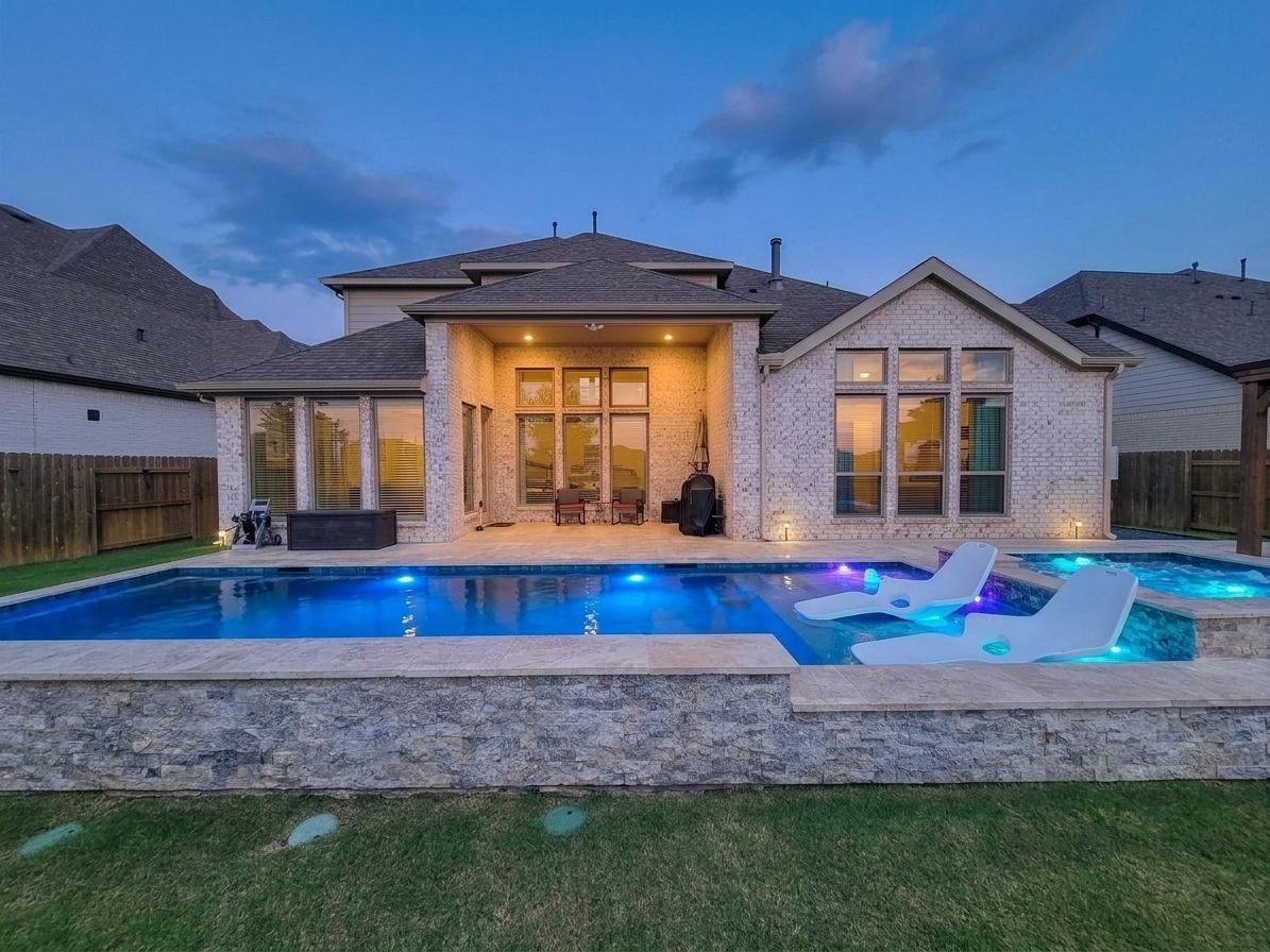 Modern home with a backyard featuring a luxurious illuminated pool and spa. The exterior is elegantly designed with large windows and a covered patio area, ideal for outdoor entertaining. Modern home with a backyard featuring a luxurious illuminated pool and spa. The exterior is elegantly designed with large windows and a covered patio area, ideal for outdoor entertaining.