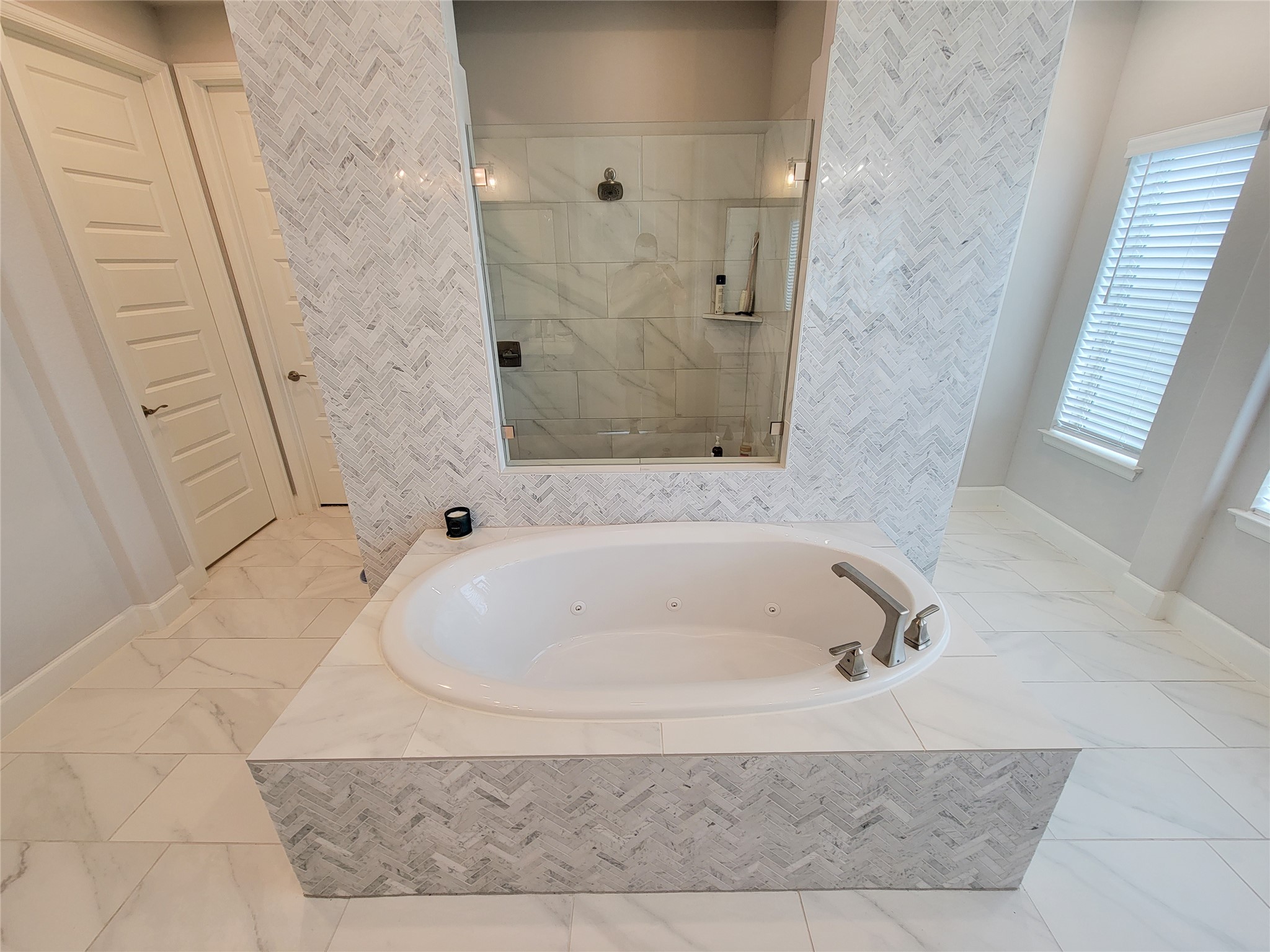 This bathroom features a luxurious soaking tub with elegant tile surround and a spacious glass-enclosed shower. The room is bright and modern, with white tiled flooring and ample natural light from the window.