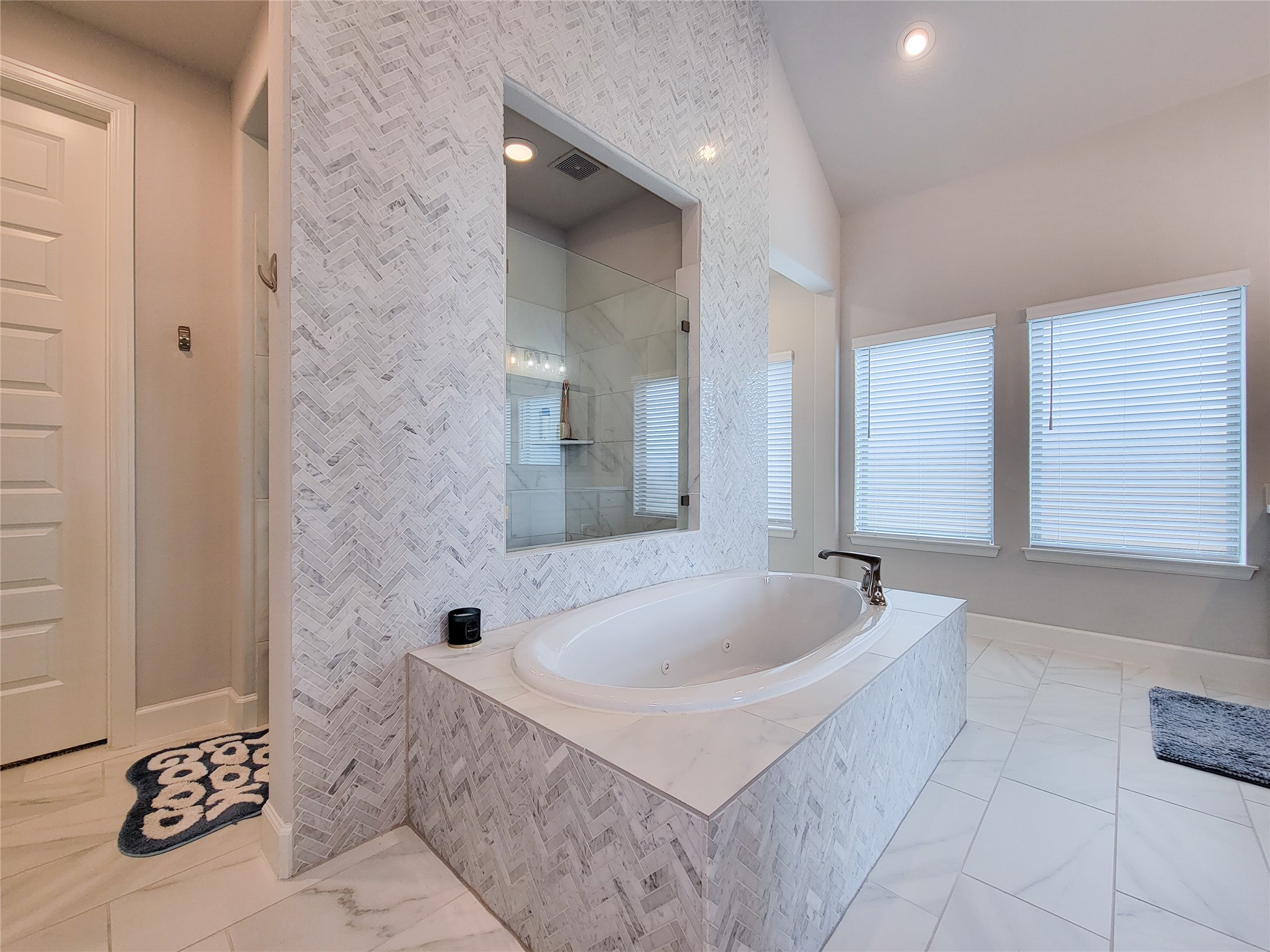 This bathroom features a luxurious, sunken tub with elegant marble tile surround and a glass-enclosed shower. The space is brightened by large windows with blinds, offering a modern and serene atmosphere.