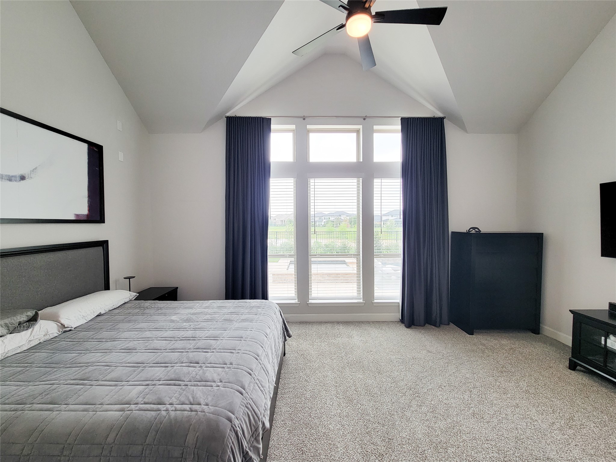 This room features a spacious layout with a vaulted ceiling and large windows, offering ample natural light. It includes a comfortable bed, modern ceiling fan, and neutral decor, providing a cozy and inviting atmosphere.