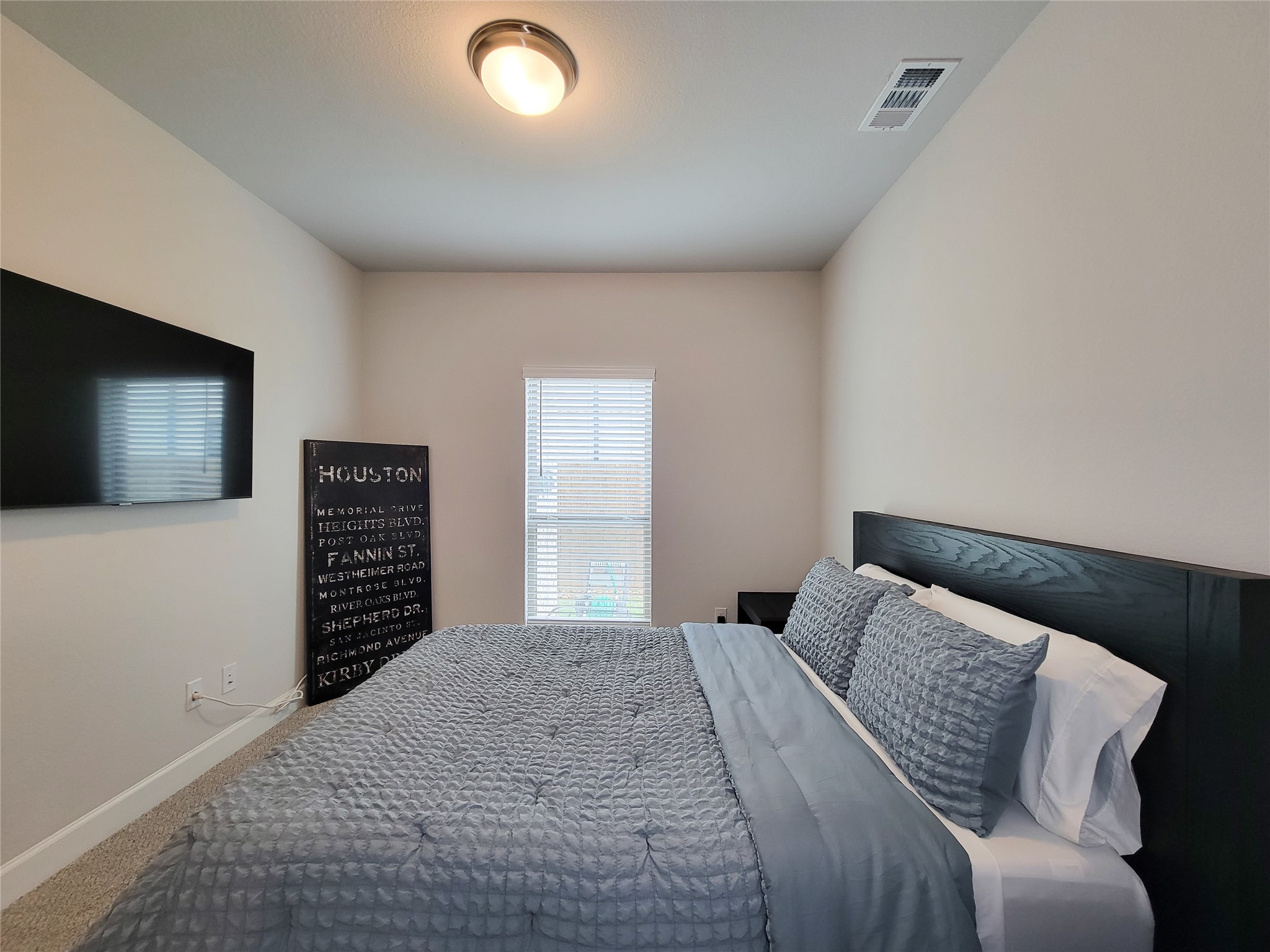This cozy bedroom features a large window allowing natural light. The room has a simple, modern design with neutral walls and a ceiling light fixture.