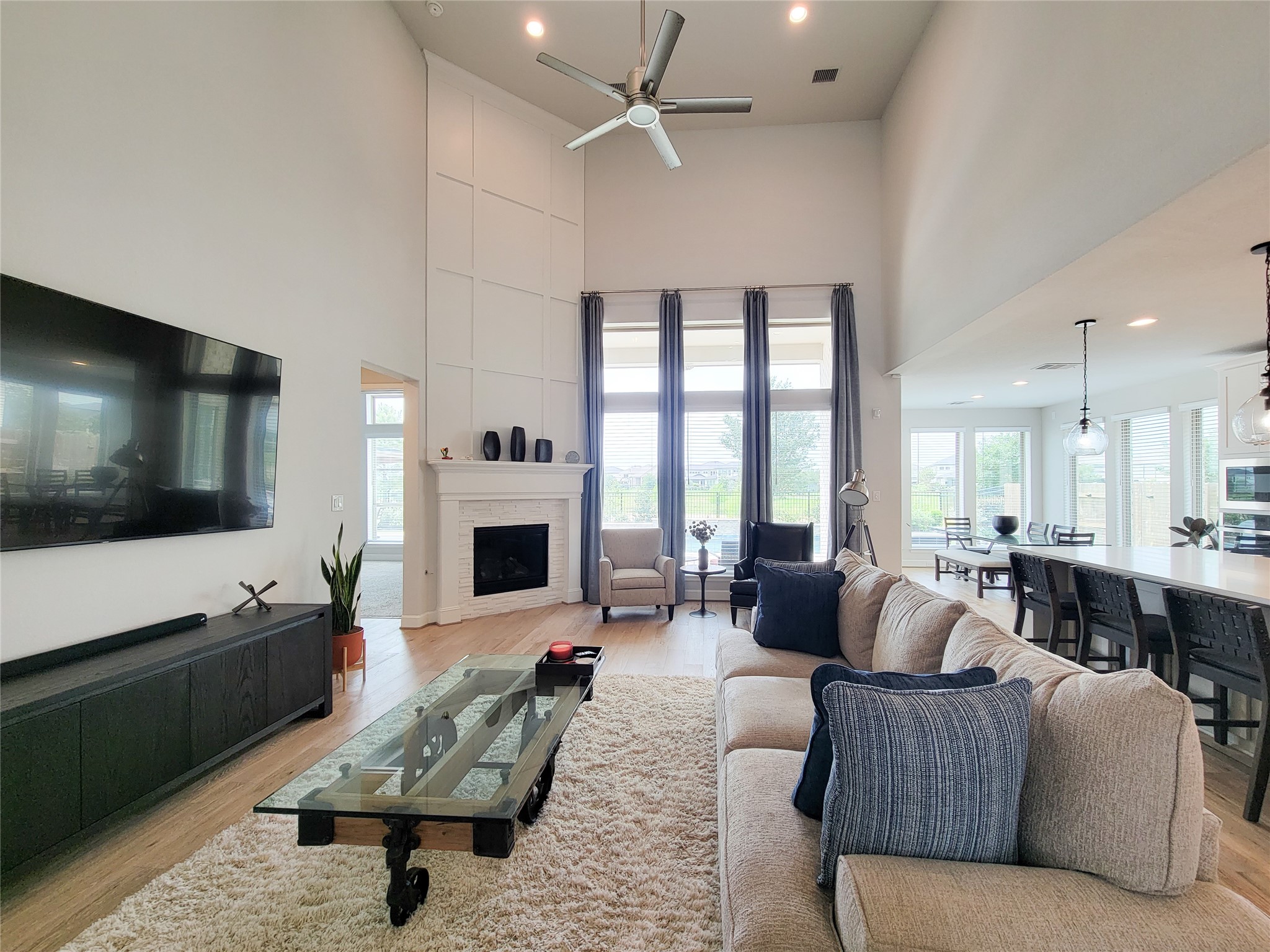 This spacious living room features high ceilings, a modern ceiling fan, and a large fireplace. Natural light pours in through tall windows, highlighting the open layout that seamlessly connects to the dining and kitchen areas.