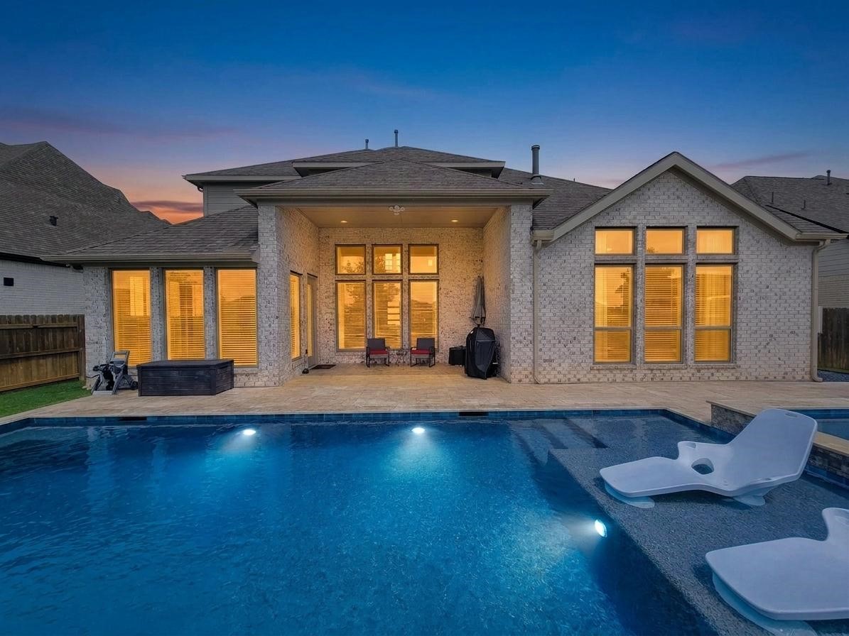 This elegant home features a spacious backyard with a beautiful illuminated pool and modern patio area, perfect for evening relaxation and entertaining. The large windows provide ample natural light to the interior.
