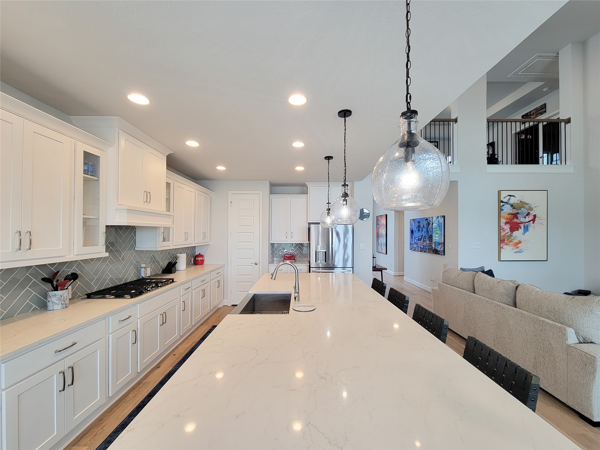 This spacious kitchen features a long marble island with pendant lighting, white cabinetry, stainless steel appliances, and an open layout leading into a living area with modern decor and high ceilings.