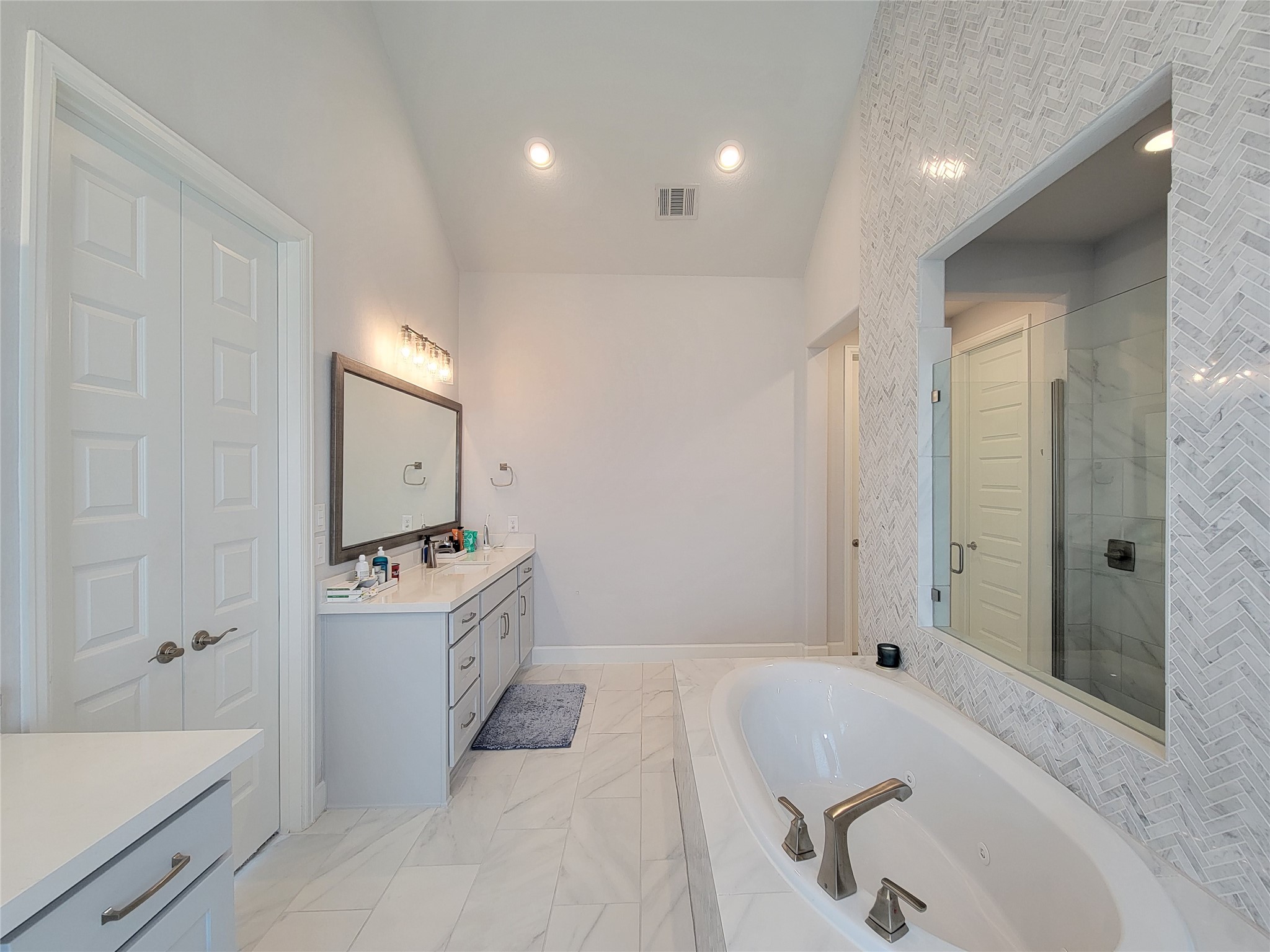 This bathroom features a modern design with a large soaking tub, spacious vanity with double sinks, and a glass-enclosed shower. The light color palette and patterned tile wall add elegance and brightness.