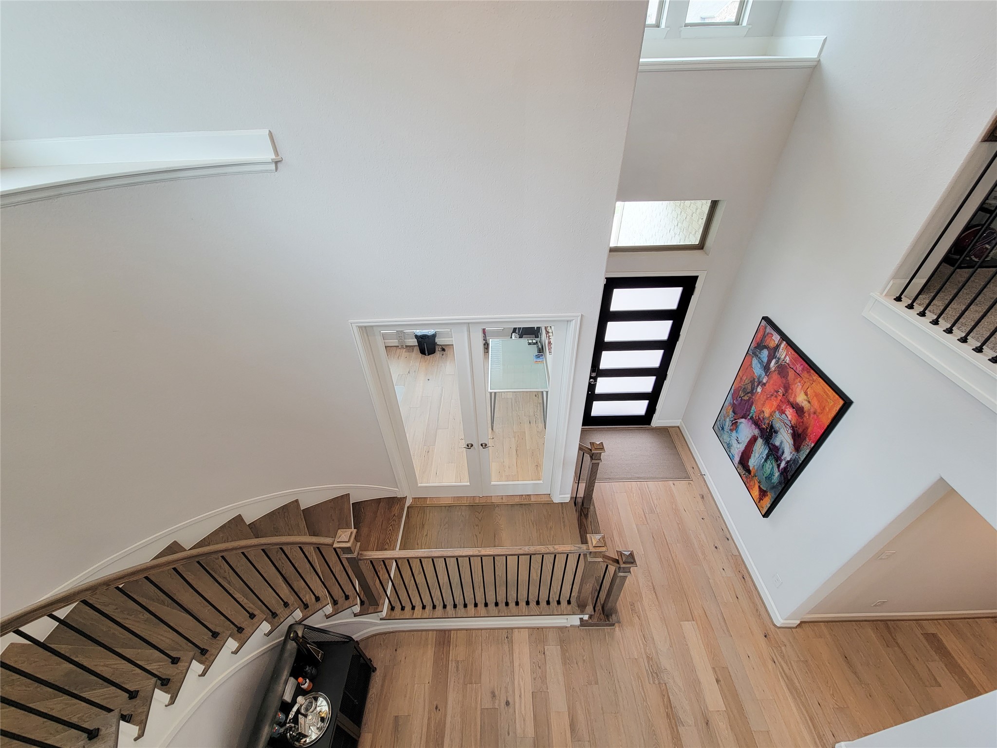 Modern, airy foyer with a curved wooden staircase and sleek black railing. The entryway features a stylish door with frosted glass panels, light wood flooring, and a colorful abstract painting, creating a welcoming and contemporary feel.