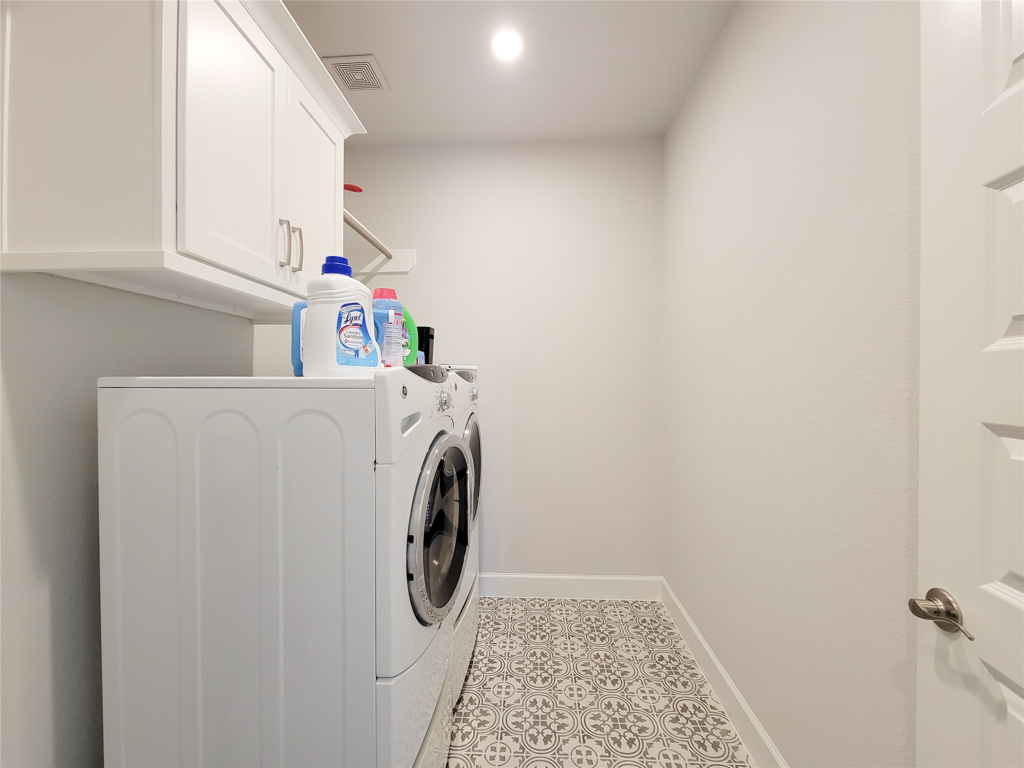 This photo shows a bright, modern laundry room featuring white cabinetry for storage and stylish patterned floor tiles.