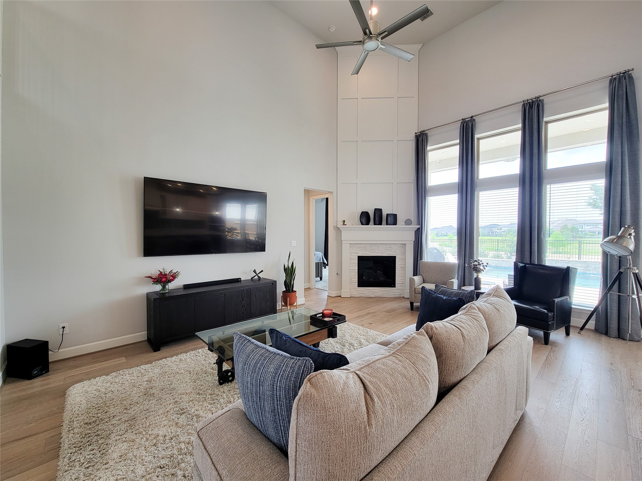This spacious living room features a high ceiling with a large ceiling fan, a cozy fireplace, and ample natural light from tall windows.