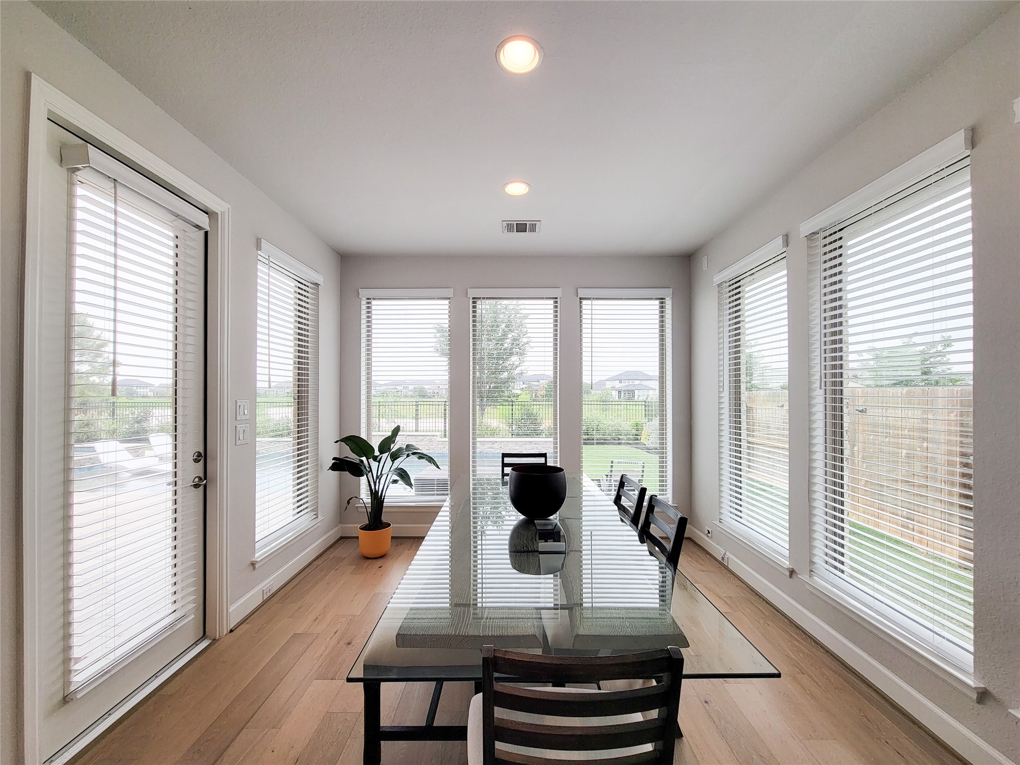This bright dining area features large windows with blinds, providing ample natural light and views of the backyard. It has a sleek glass table, modern chairs, and wood flooring, creating an inviting space for meals and gatherings.