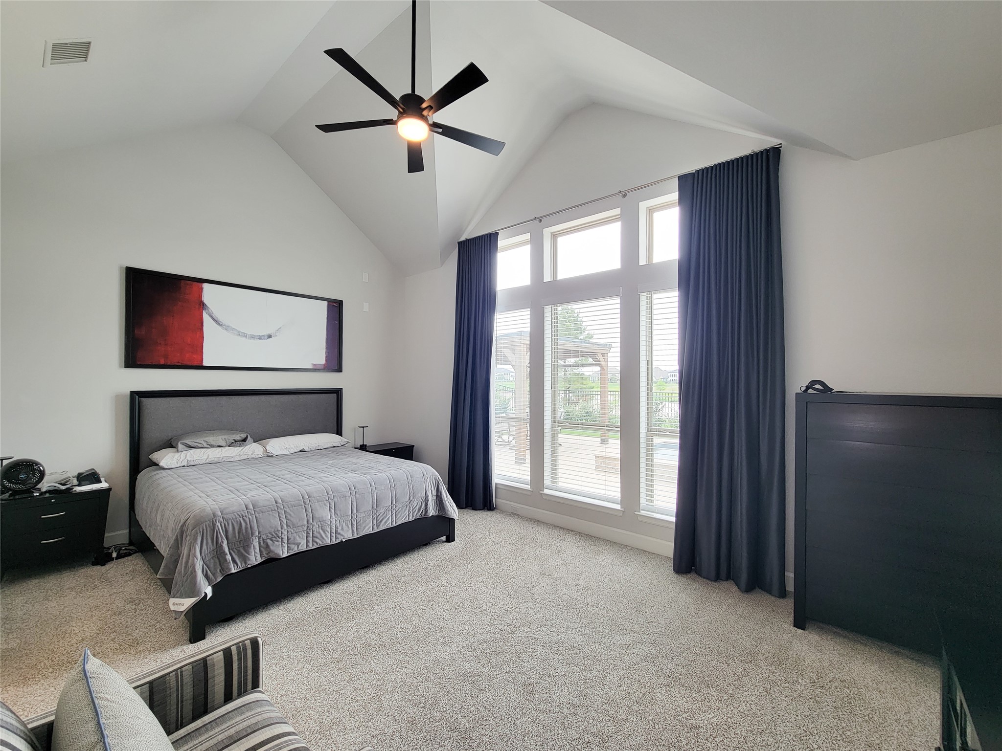 This spacious bedroom features a high vaulted ceiling with a modern ceiling fan, large windows with elegant curtains, and ample natural light. It includes a comfortable bed, contemporary furniture, and neutral carpeting, creating a cozy and inviting atmosphere.