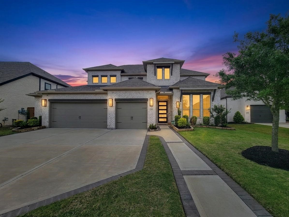 This modern two-story home features a spacious three-car garage and a welcoming walkway. It is beautifully lit against a stunning sunset backdrop, with well-maintained landscaping and contemporary architectural design.