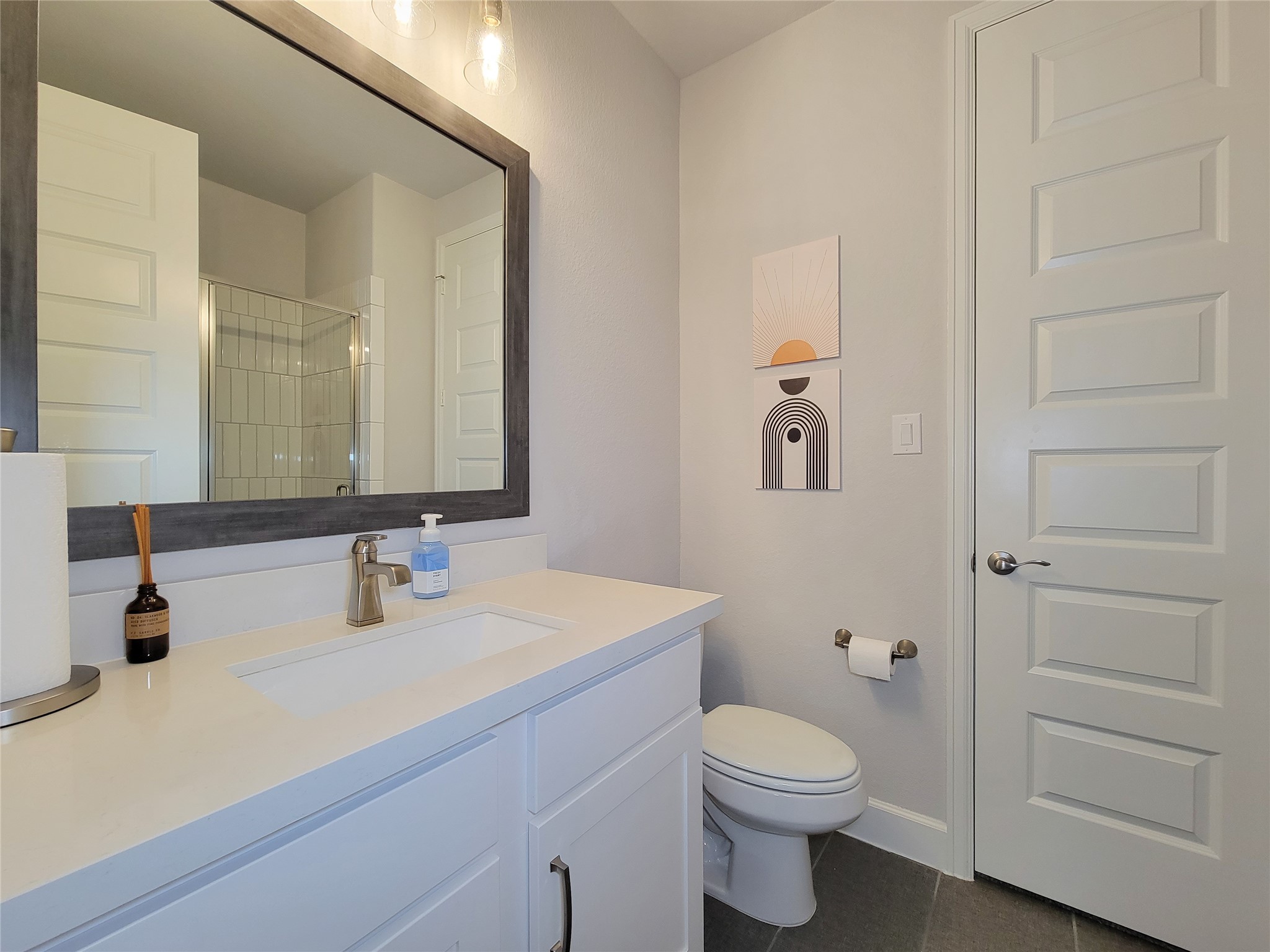 This bathroom features a sleek, modern design with a large mirror above a white vanity, neutral tones, stylish wall art, and a spacious shower with glass doors.