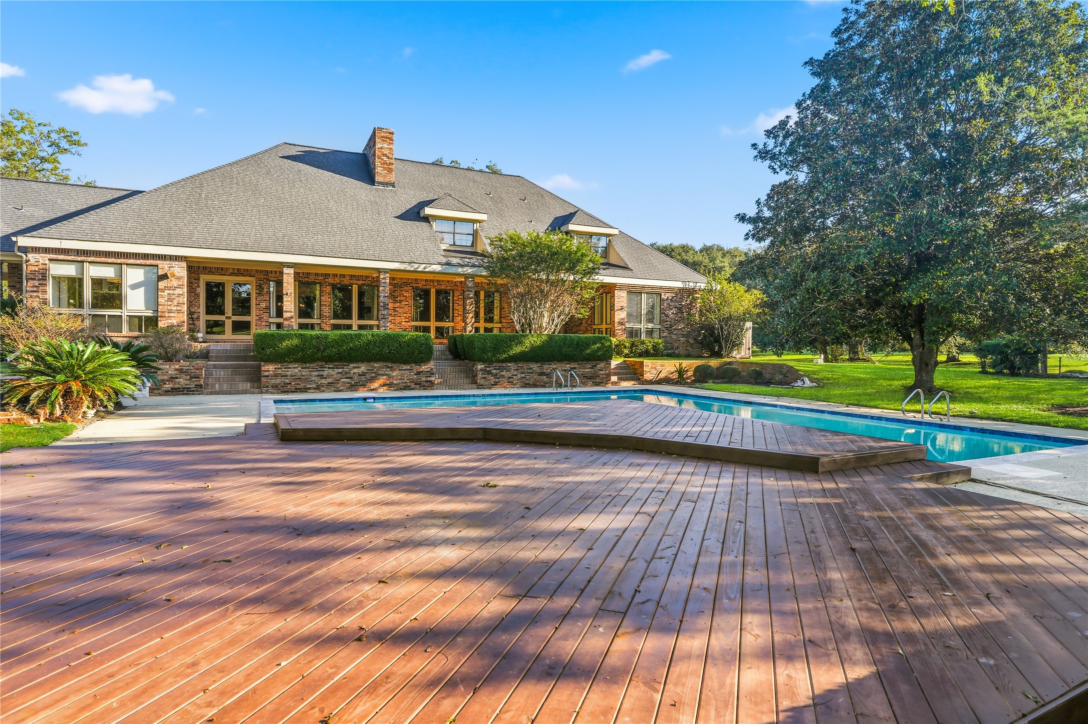 Charming brick home with expansive windows opens to a spacious wooden deck and inviting swimming pool, surrounded by lush greenery and mature trees.